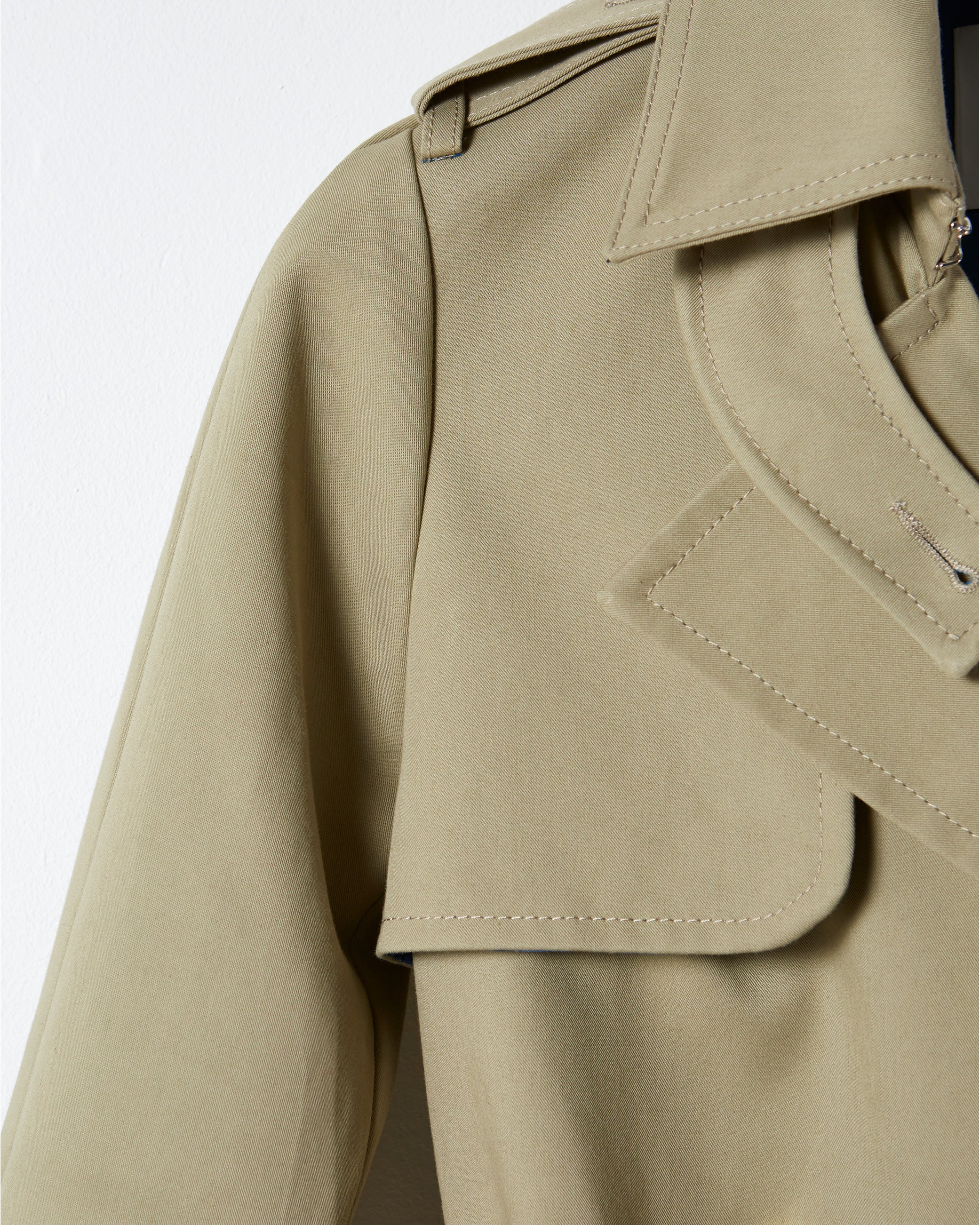 Close-up of a beige trench coat showing the collar, lapel, and shoulder detail against a white background.