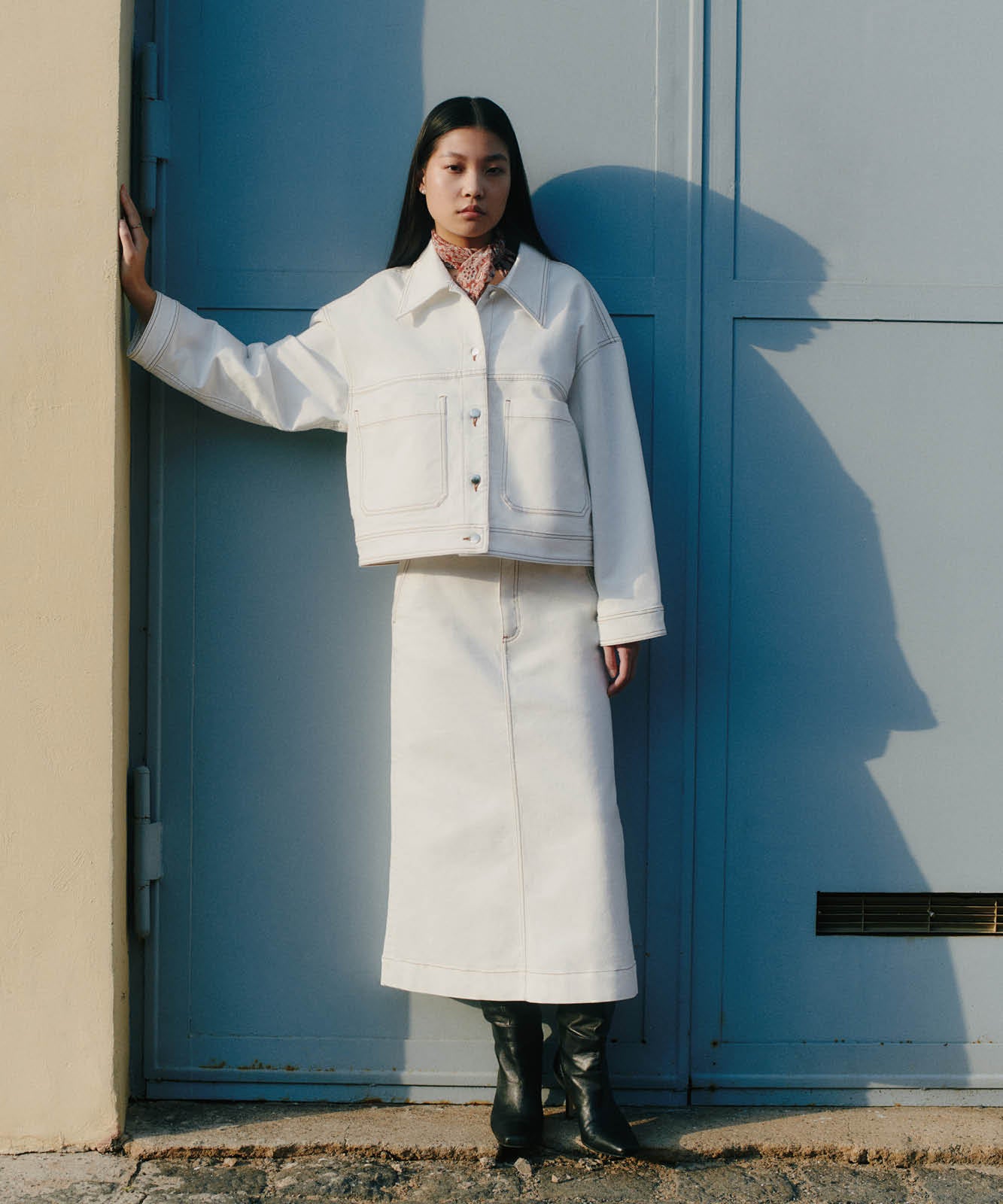 Woman in a white jacket and skirt, black boots, standing against a blue door, hand on wall.