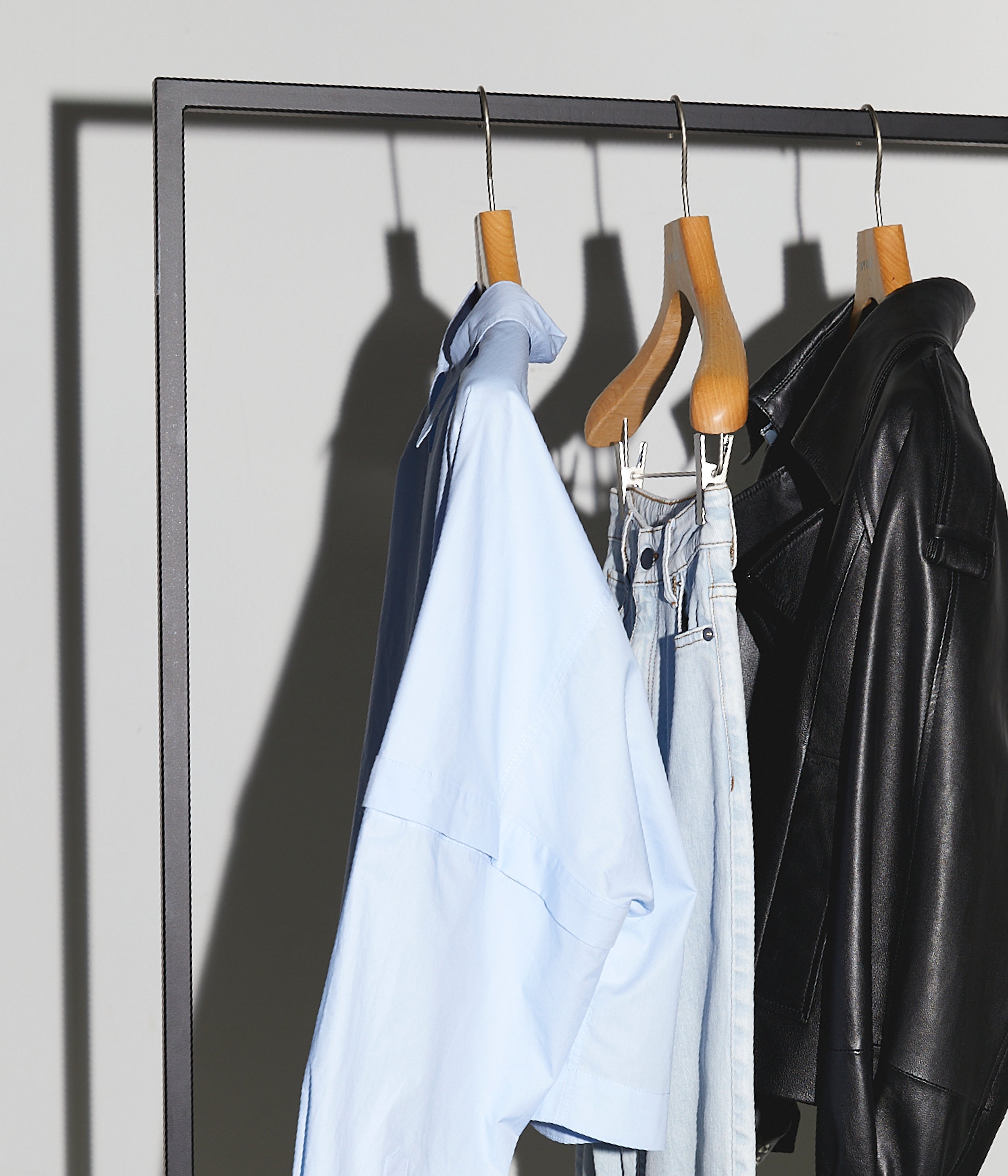 Light blue shirt, light denim jeans, and black leather jacket hanging on wooden hangers on a metal rack.