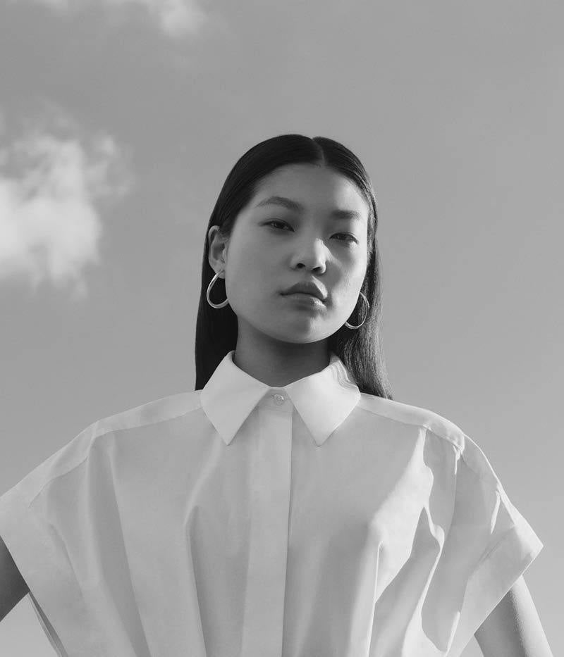 Woman with long hair in a white shirt and hoop earrings, looking serious against a cloudy sky. Black and white photo.