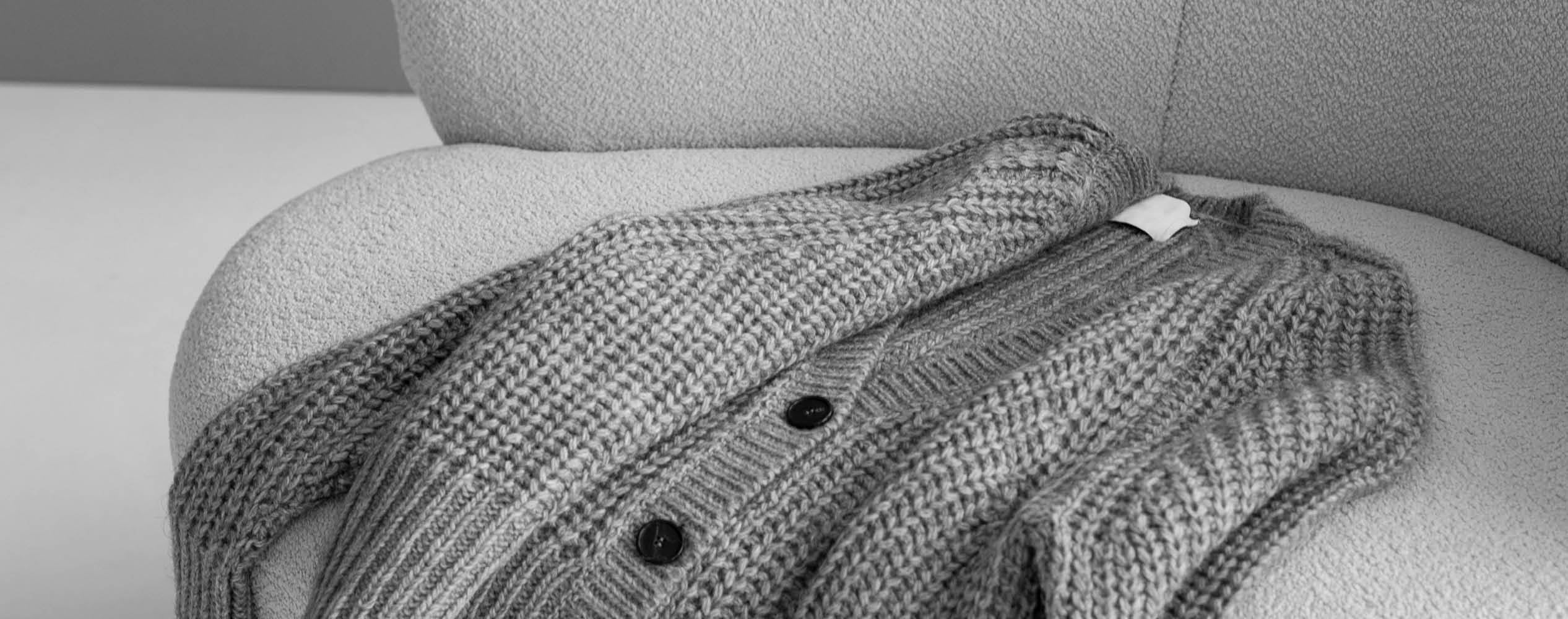 A chunky knit cardigan with dark buttons draped over a textured, light-colored sofa.