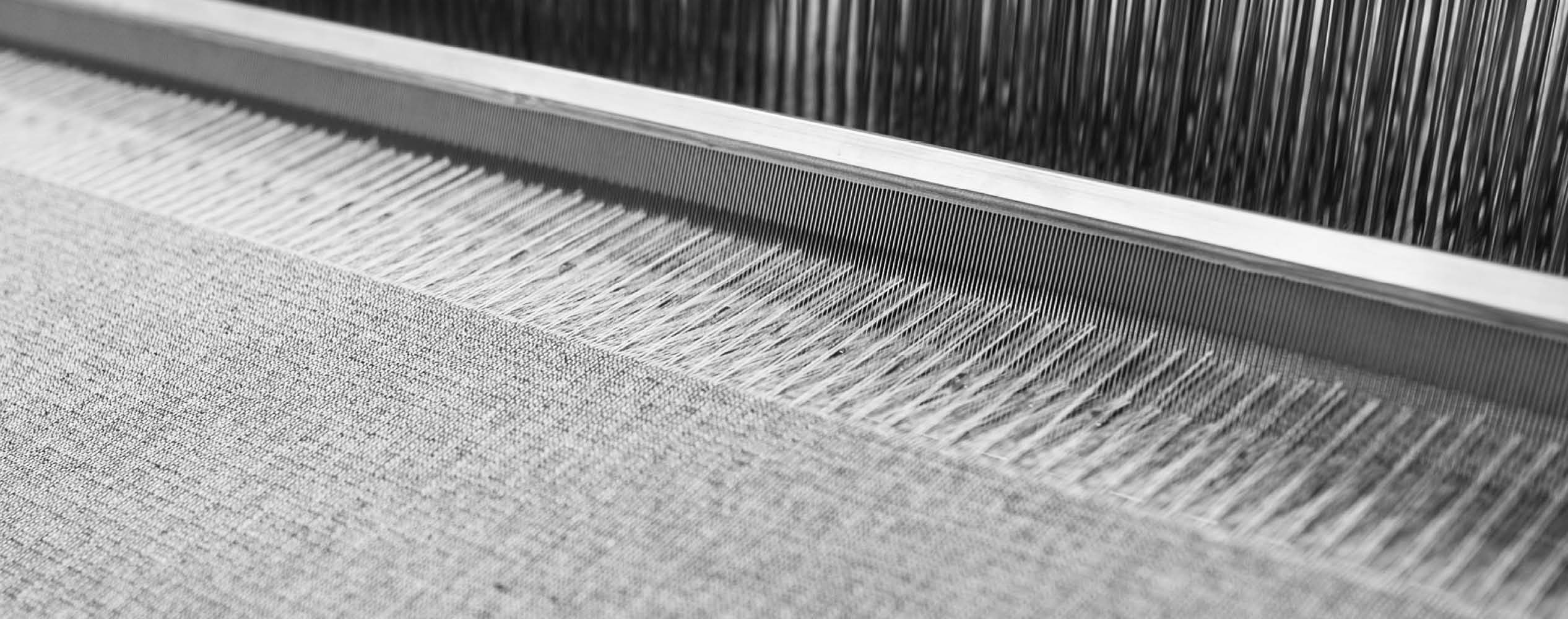 Close-up of threads being woven on a loom, forming fabric in black and white.