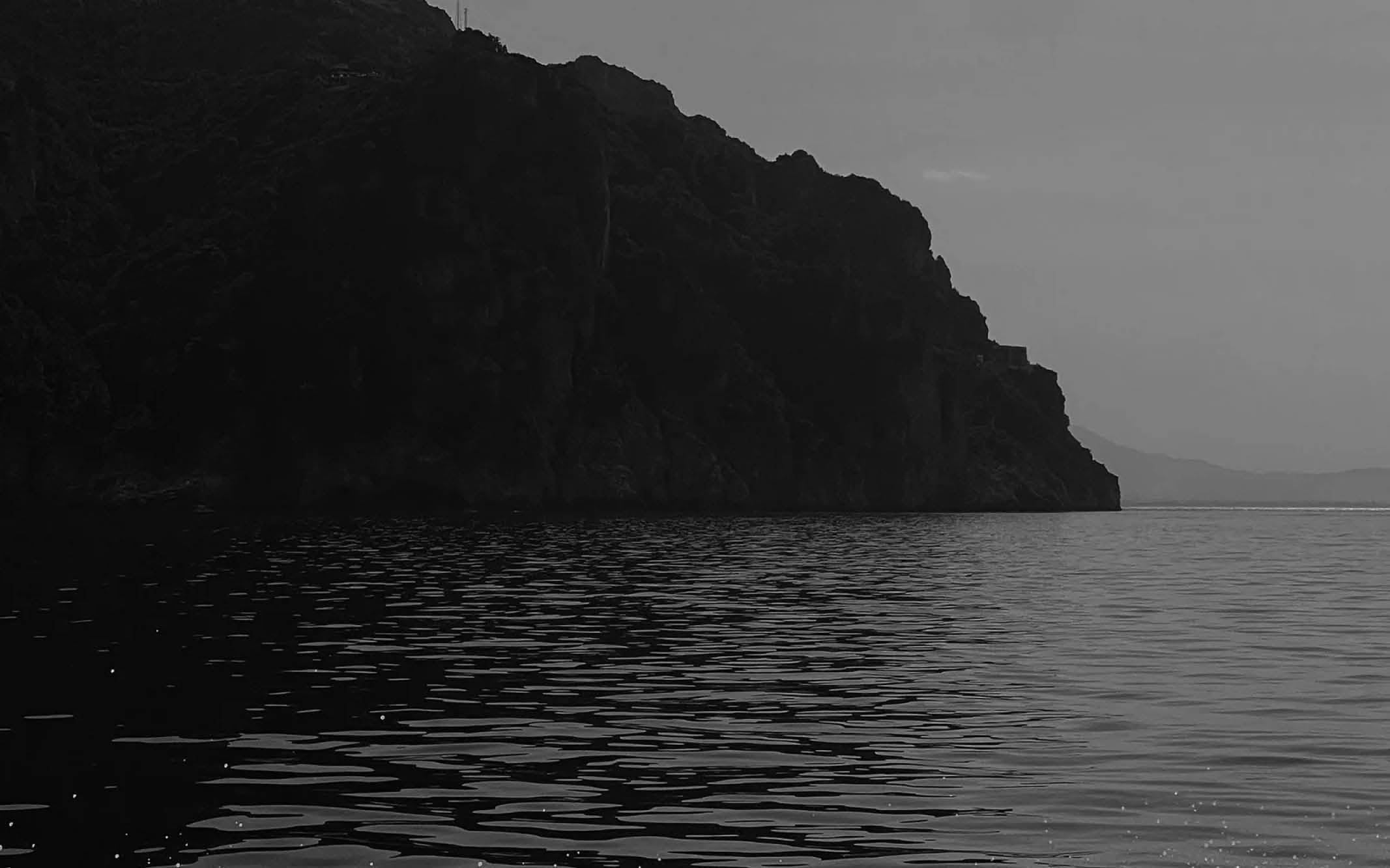 Rocky cliff by calm water under a cloudy sky, captured in black and white.