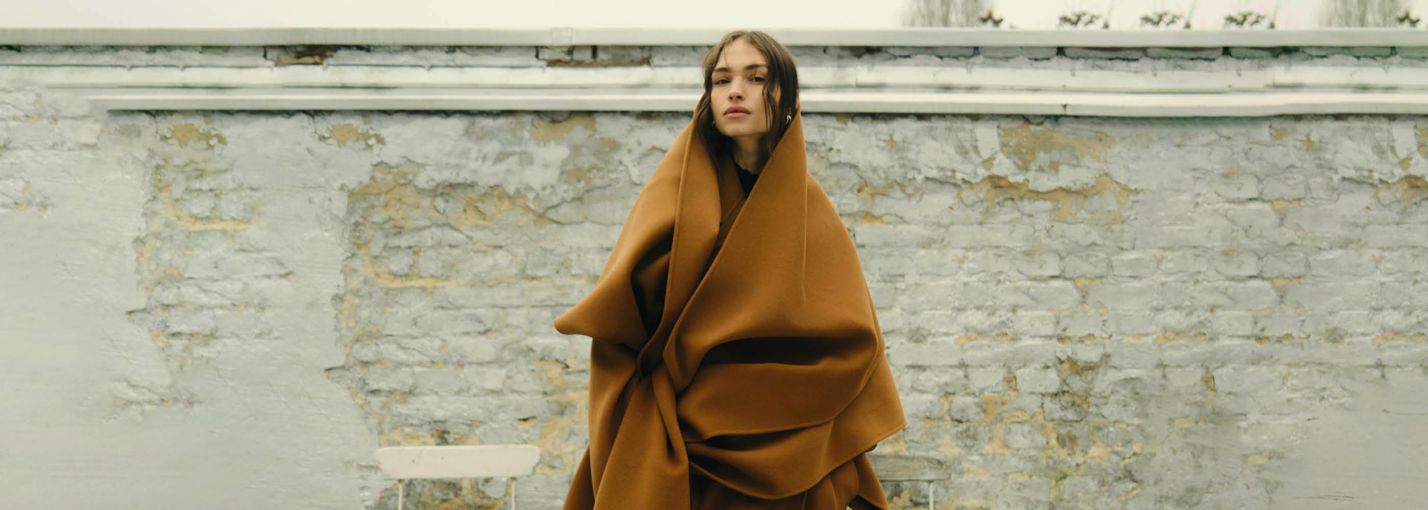 Person wrapped in a large brown blanket stands in front of a weathered, light-colored brick wall outdoors.