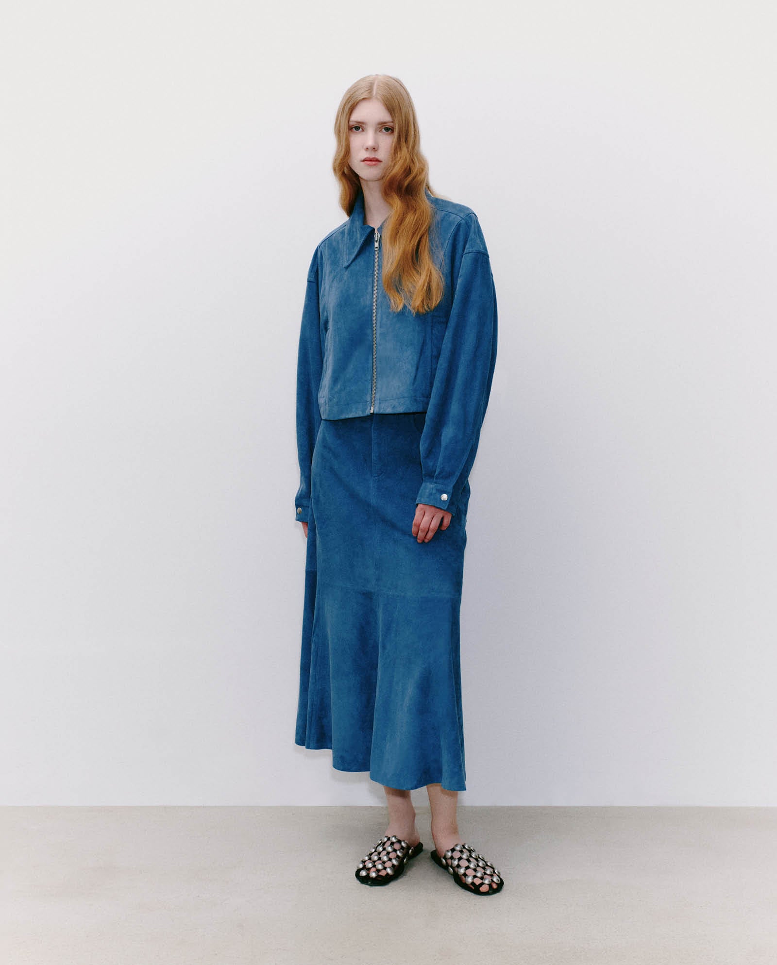 A woman stands against a plain white wall wearing the IVY OAK LENA Jacket—a blue, cropped, boxy suede bomber—paired with a skirt and black-and-white shoes.