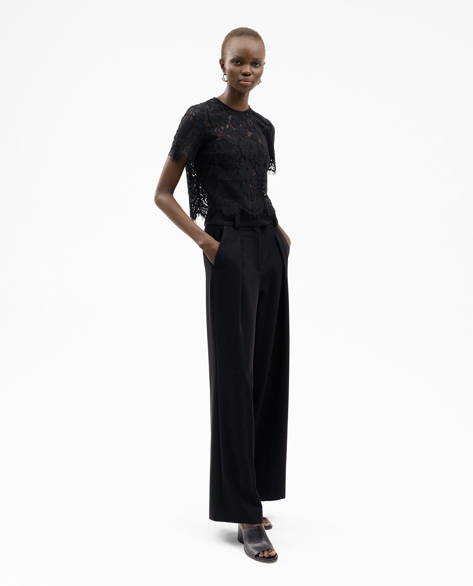 Wearing the IVY OAK TRACEY Top, a person stands with hands in pockets, styled with wide-leg black pants and open-toe heels.