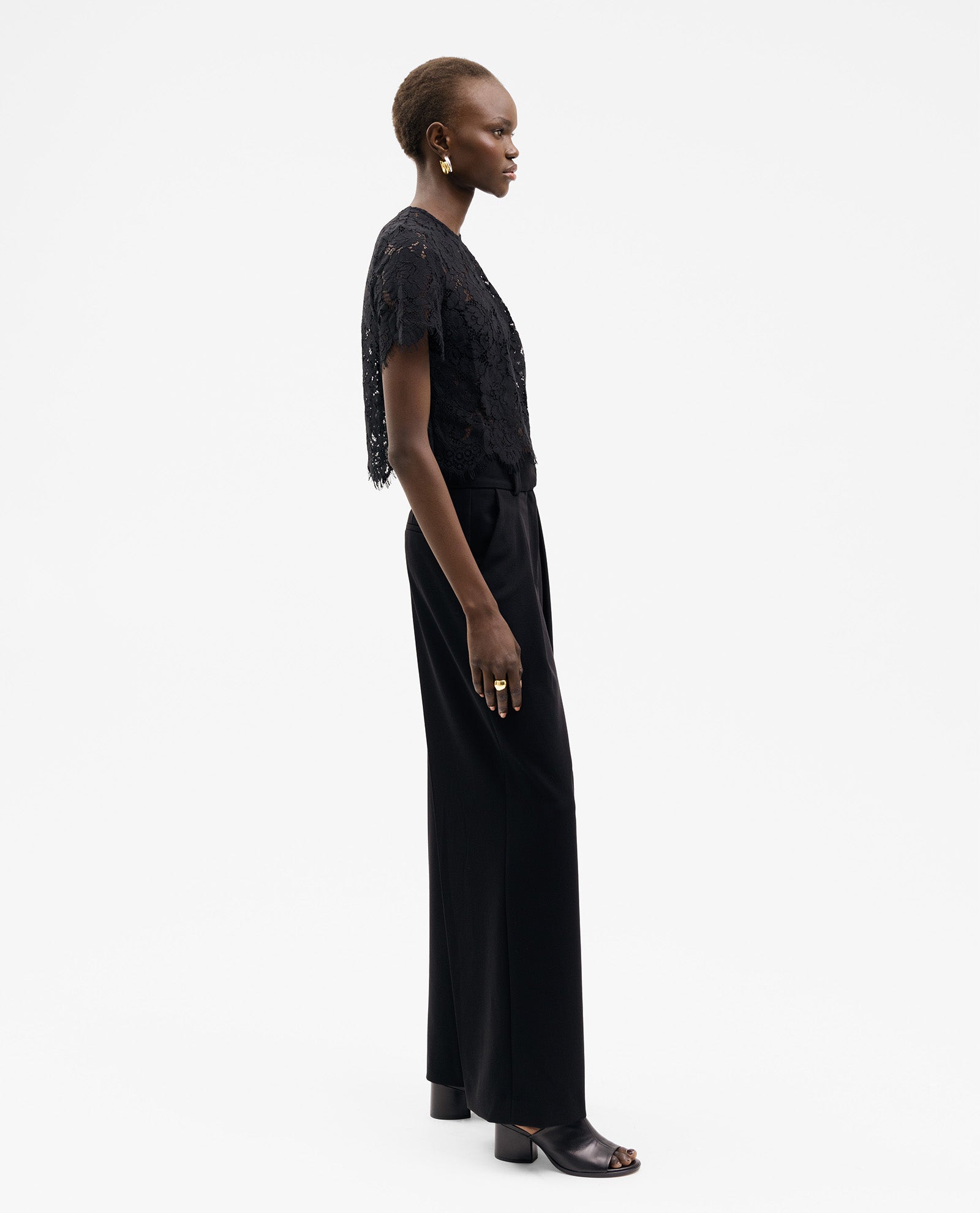 Woman in profile wears the TRACEY Top by IVY OAK, a black lace short-sleeve piece with a boxy fit, styled with wide-leg black pants and open-toe shoes, set against a white background.