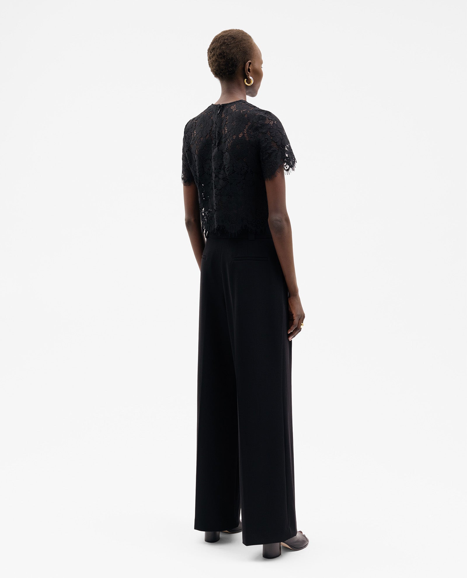 Model with short hair wears the IVY OAK TRACEY Top, a black lace, short-sleeve boxy-fit blouse, paired with wide-leg pants, standing and facing away.