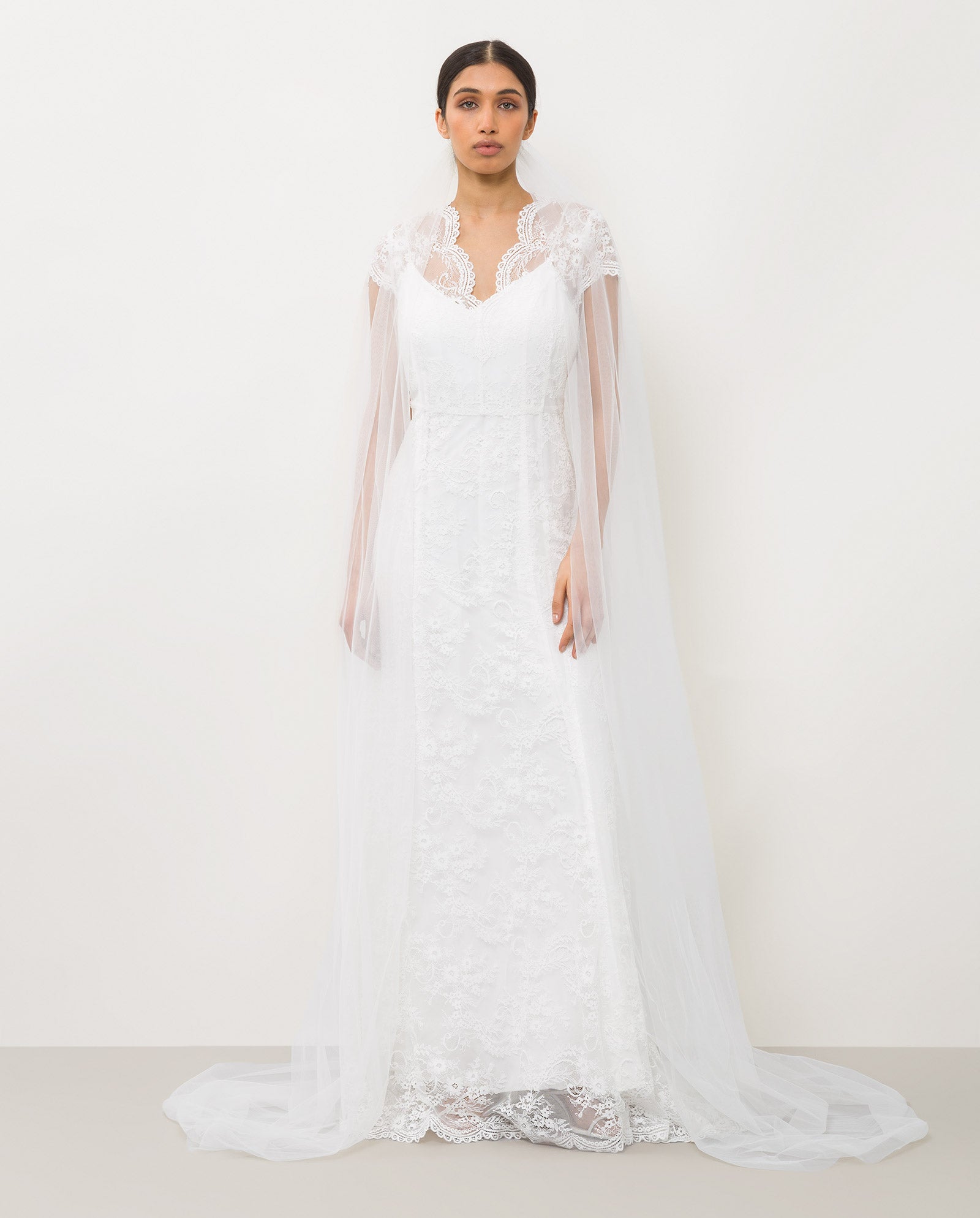 A woman wears the APHRODITE Veil by IVY OAK with a long, white lace wedding dress featuring sheer sleeves and a lace-trimmed neckline.