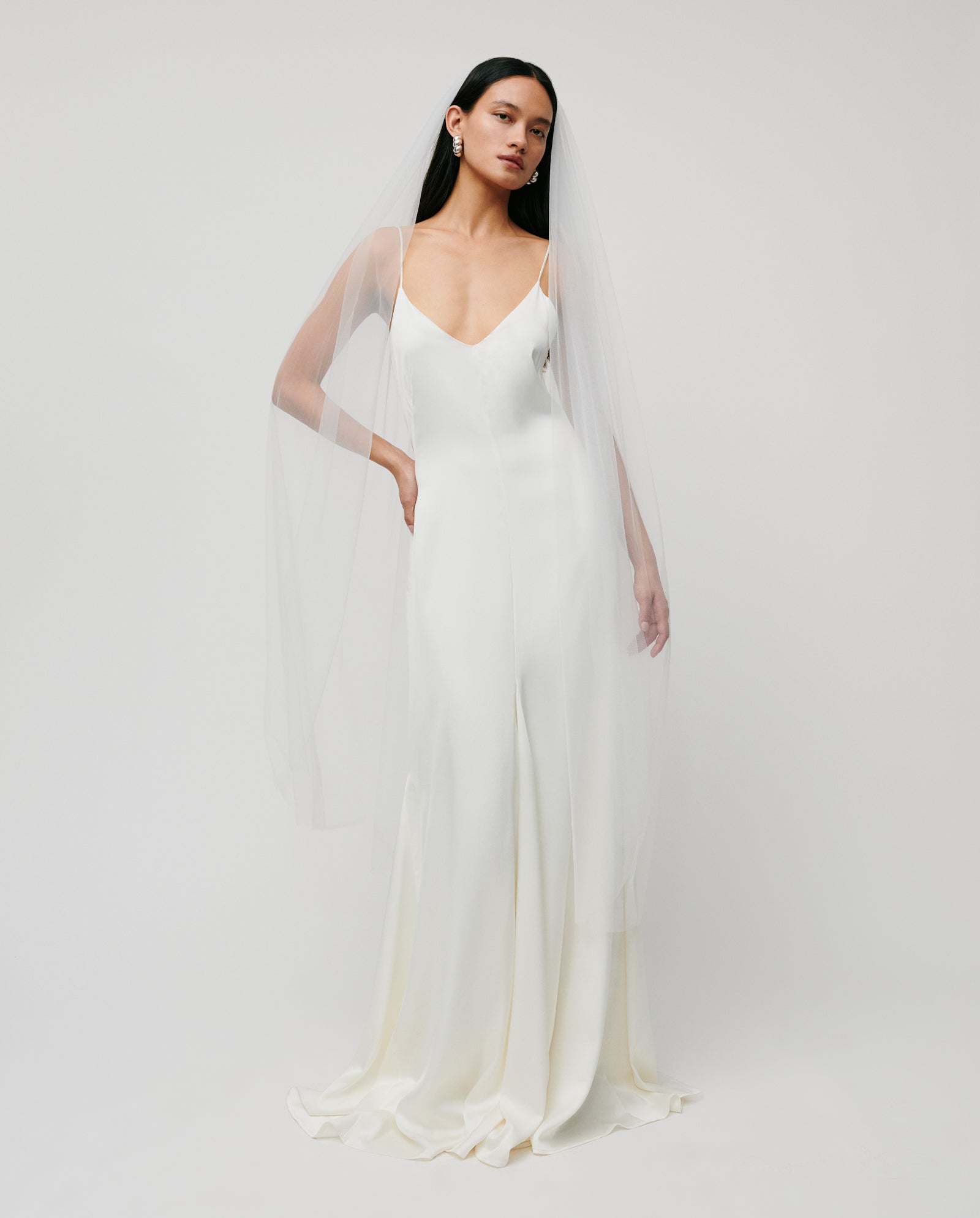 A woman stands against a plain background wearing a simple white wedding dress, adorned with the delicate APHRODITE ROSE Veil by IVY OAK.