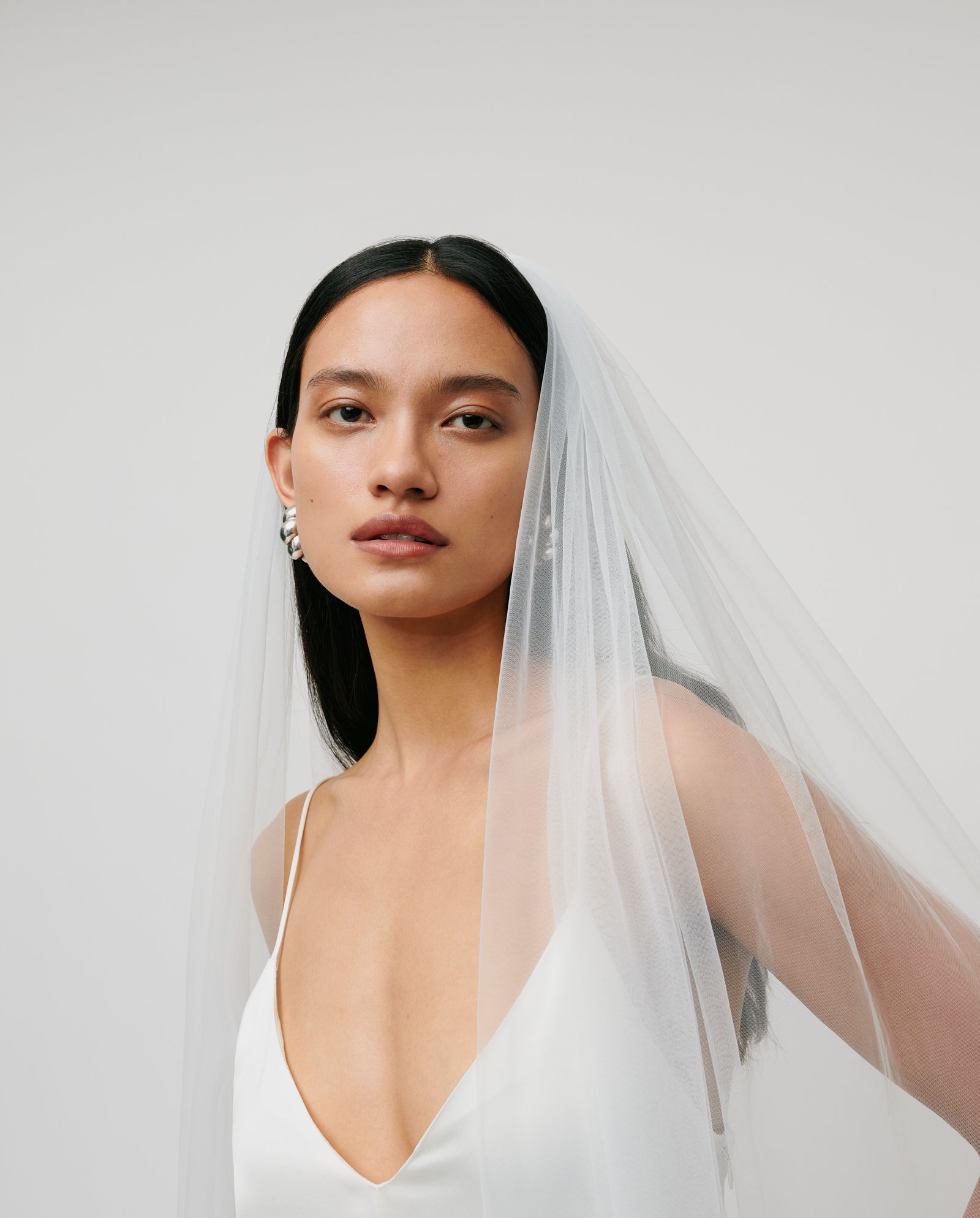 A woman wears the IVY OAK APHRODITE ROSE Veil with a white dress, gazing at the camera against a simple light background.