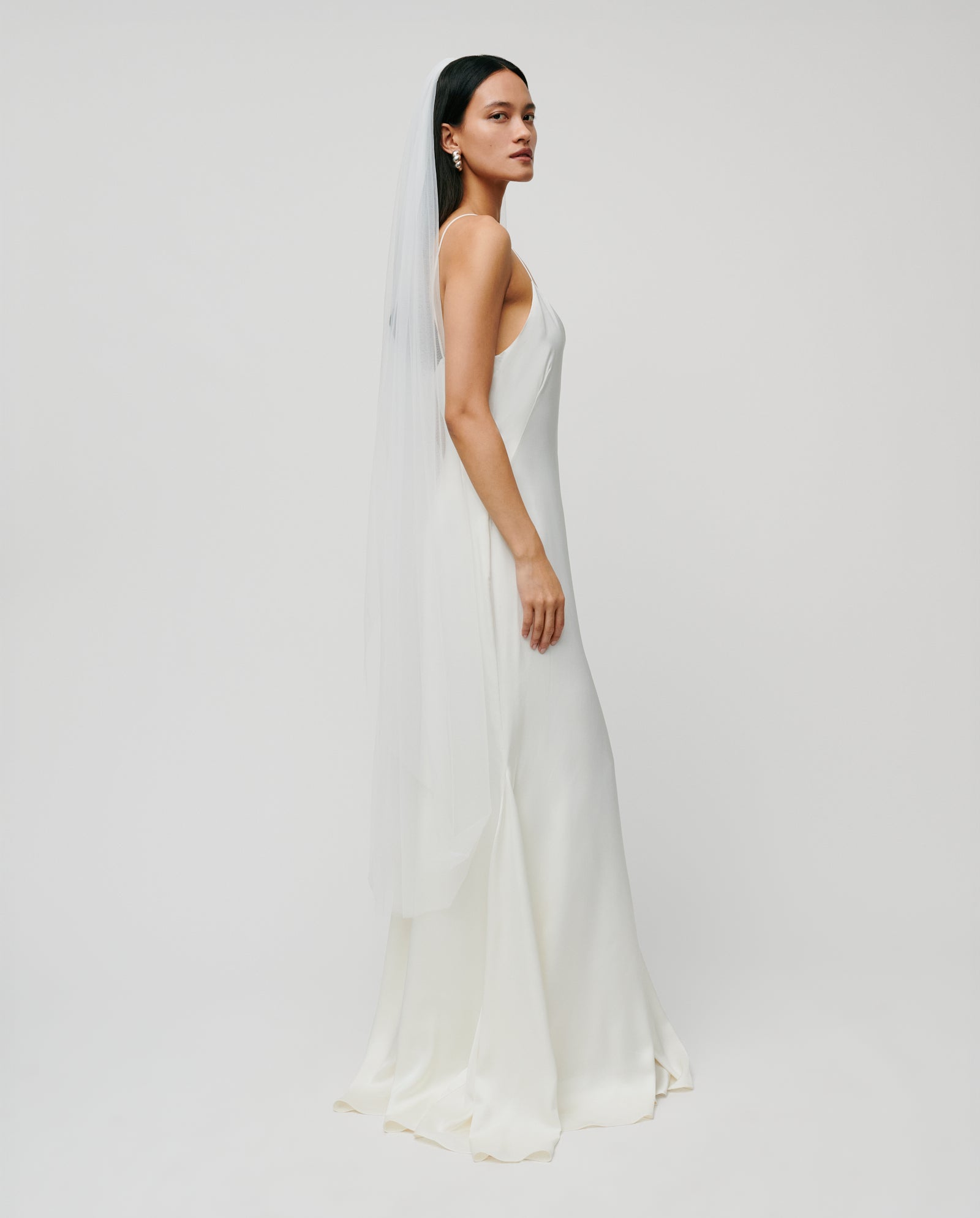 A woman in a simple white sleeveless wedding dress wears the IVY OAK APHRODITE ROSE Veil, featuring a long, delicate lace-edged design, standing against a plain background.