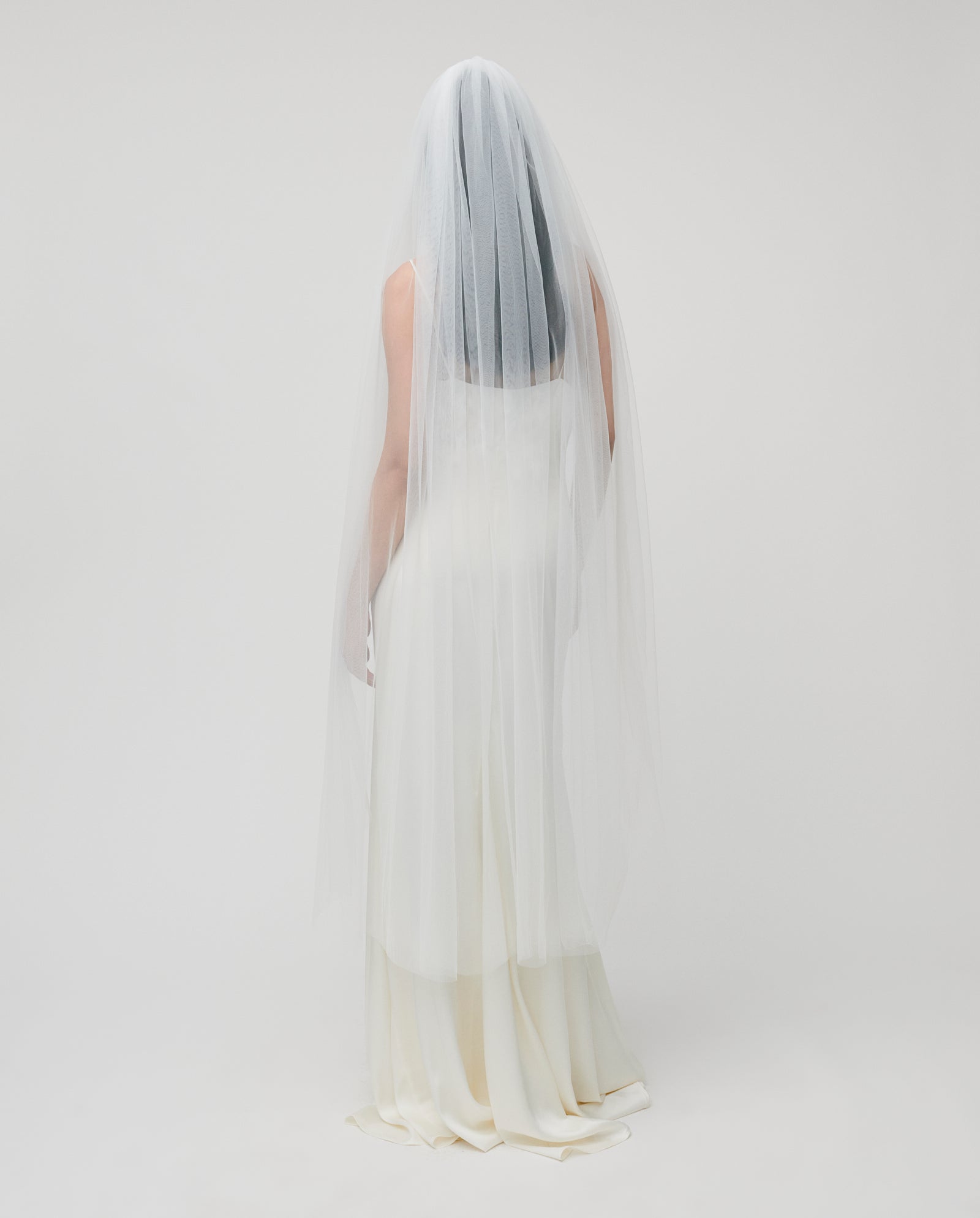 A bride wearing the IVY OAK APHRODITE ROSE Veil and a long white gown stands facing away against a plain, light background.