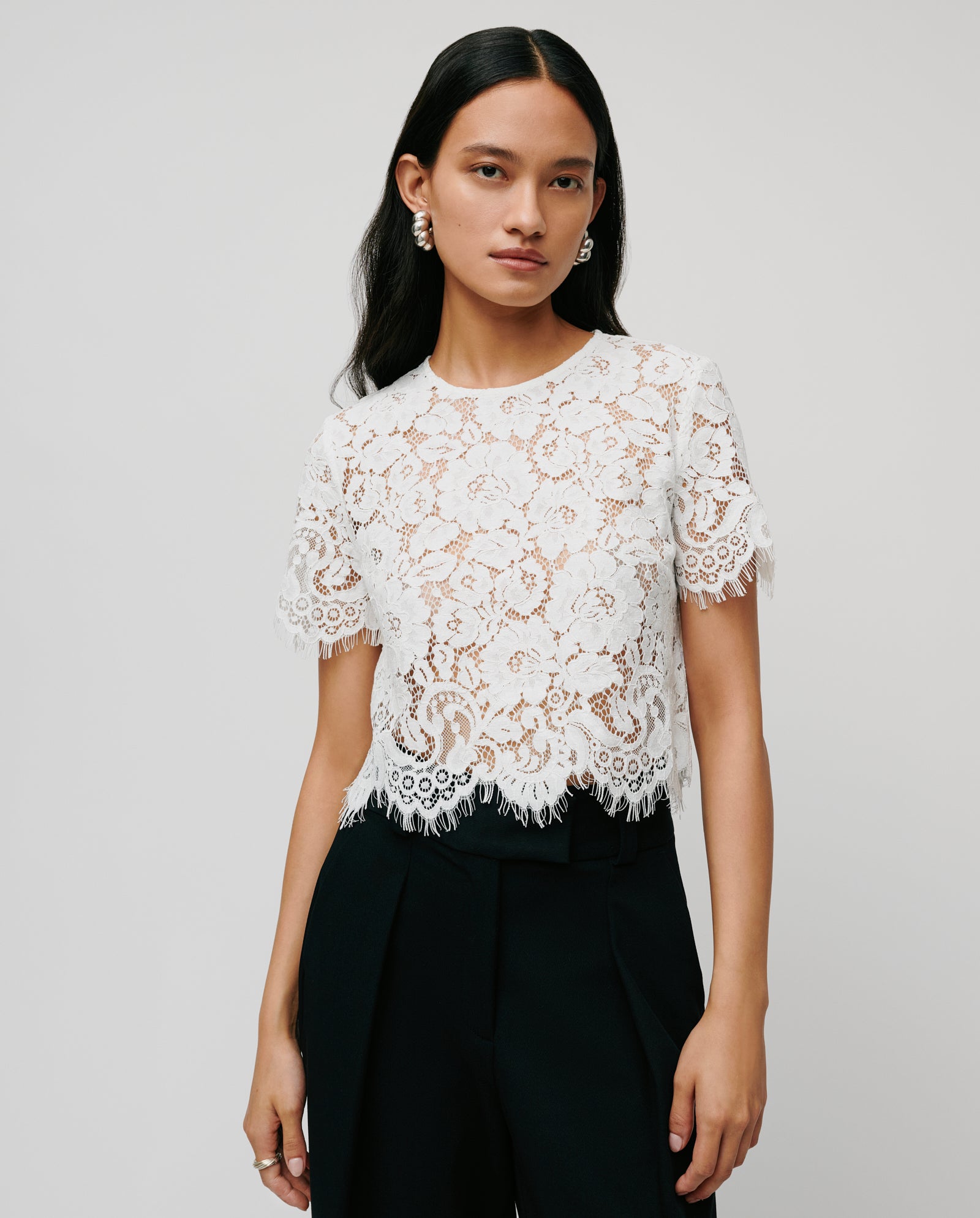 Woman wearing the TRACEY Top by IVY OAK, a white lace short-sleeve blouse aus feiner Viskose, paired with black pants and standing against a plain background.