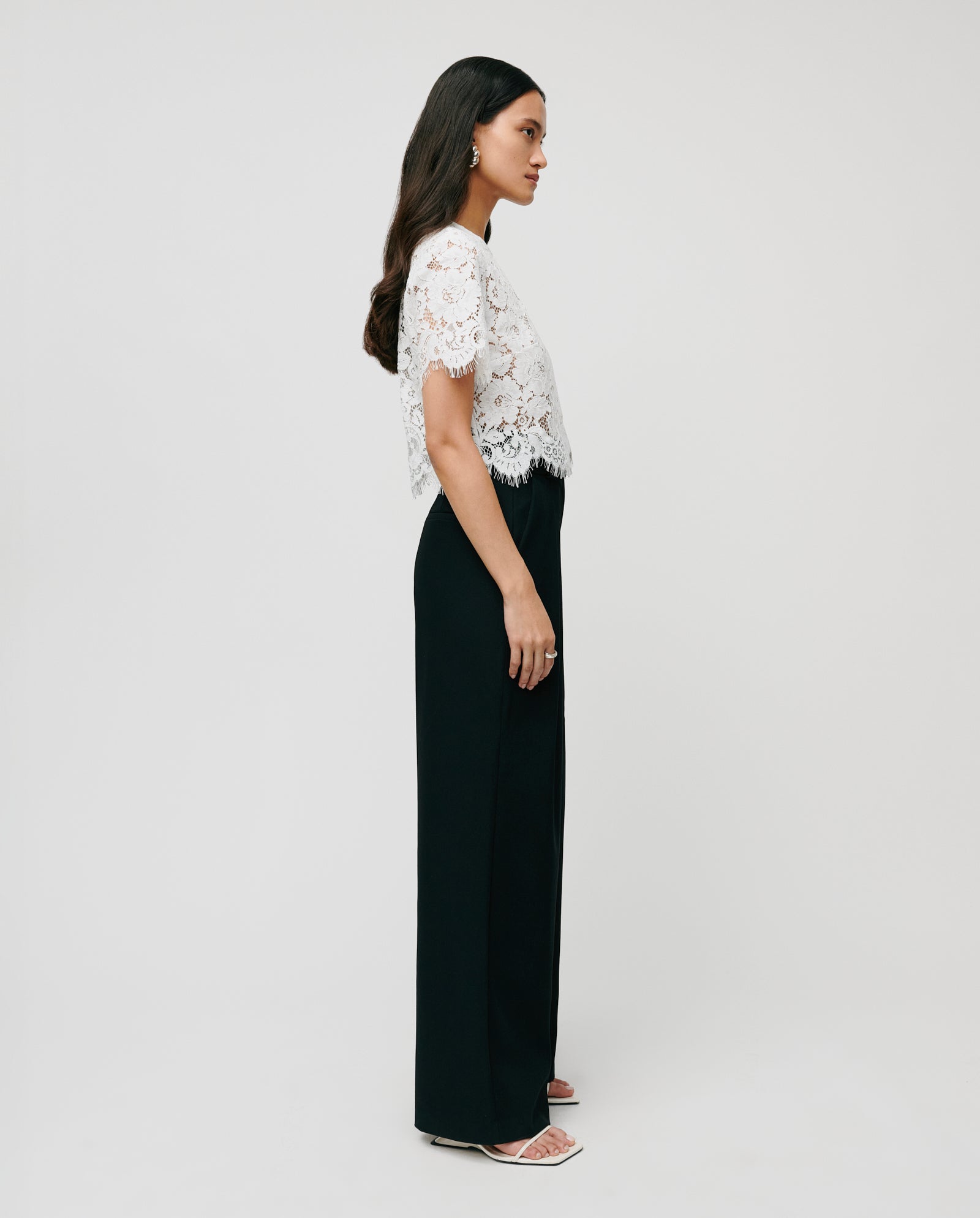 A woman in profile wears the IVY OAK TRACEY Top, a white lace piece with short sleeves and relaxed fit, styled with black high-waisted pants and white heeled sandals.