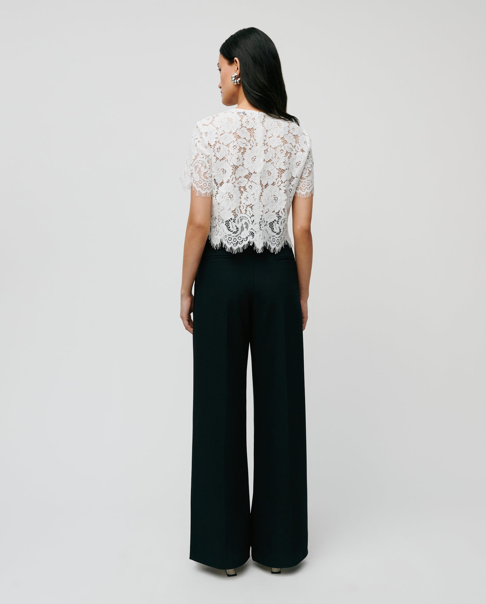 A woman faces away, wearing the IVY OAK TRACEY Top in white lace with kurzen Ärmeln and high-waisted black wide-leg pants in feine Viskose, presenting a lockere Passform against a plain background.
