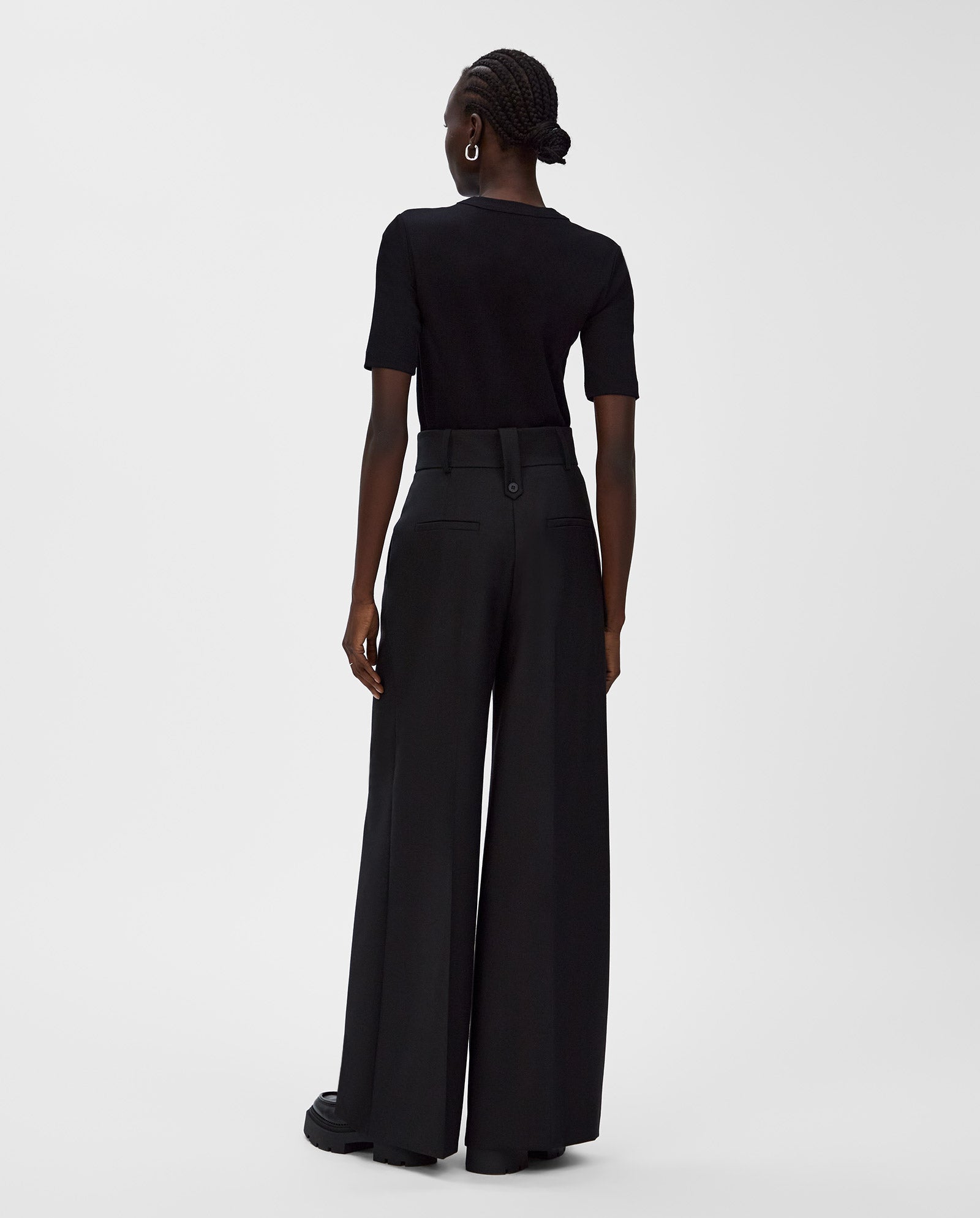 A woman stands with her back to the camera, wearing an IVY OAK KRISTIN T-Shirt in black paired with wide-leg black pants.