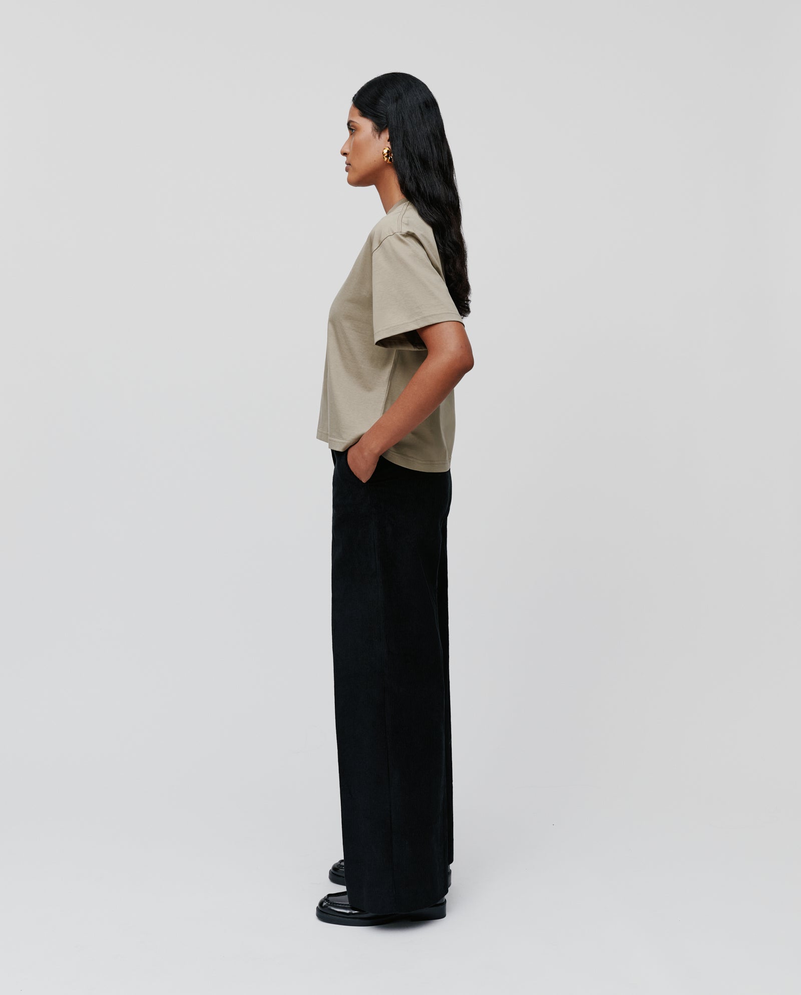 A woman stands in profile wearing the IVY OAK KYLA Top made from organic cotton and black wide-leg pants against a plain background.