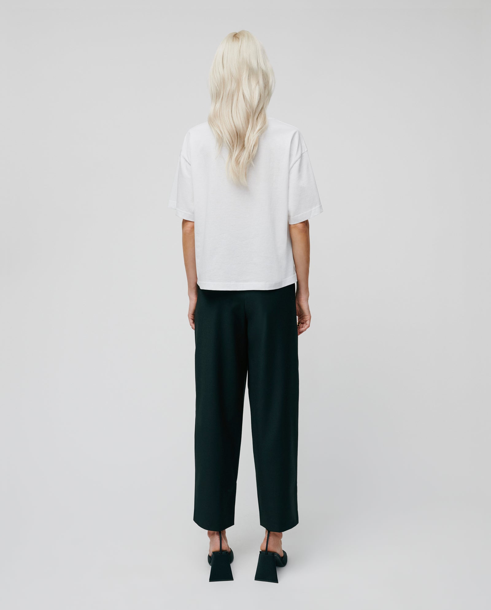 Person with long blonde hair wears the IVY OAK KYLA Top, made from organic cotton, paired with black pants and stands facing away.