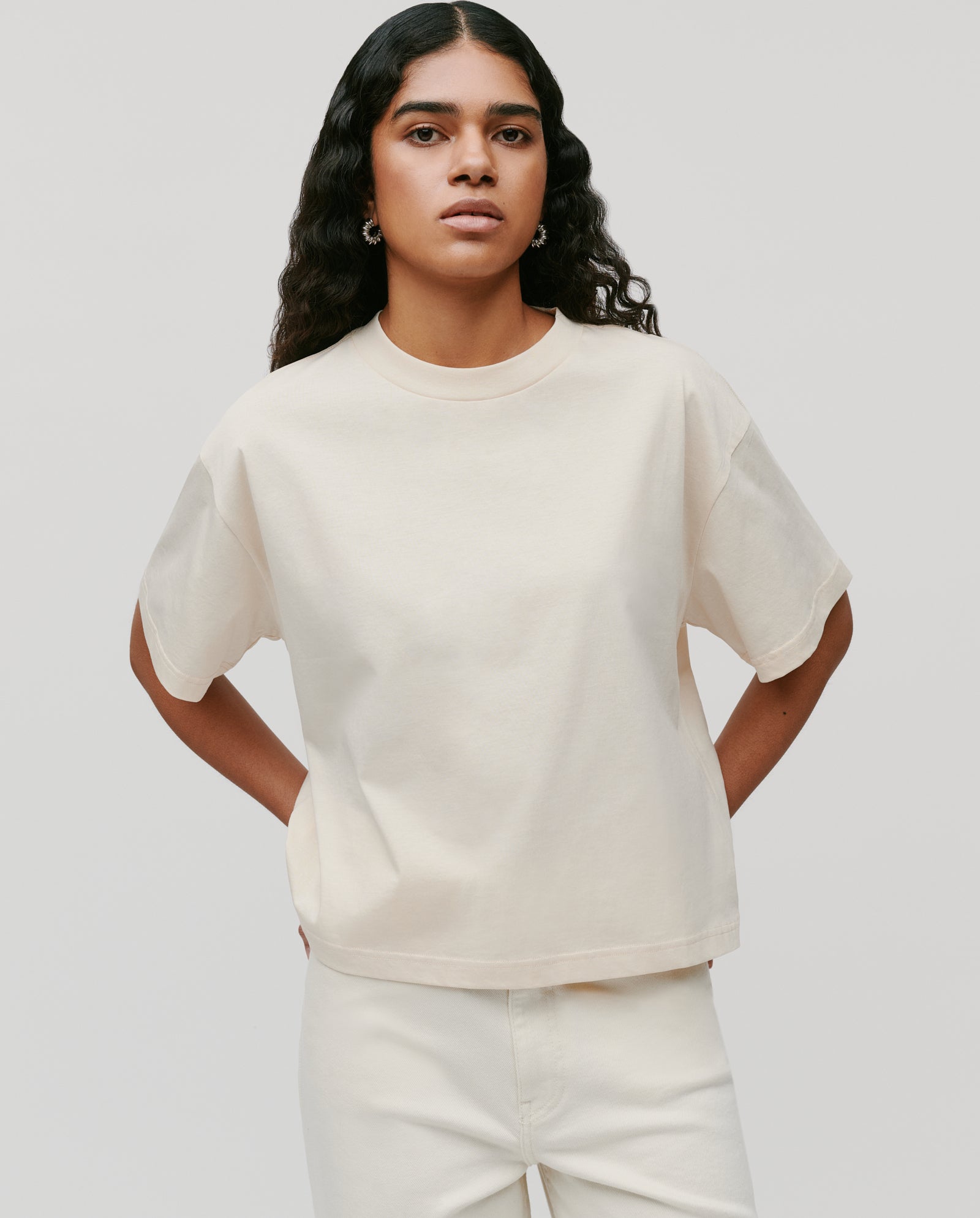 Woman wearing the IVY OAK KYLA Top, a loose, light beige boxy T-shirt in organic cotton, paired with matching pants—an effortless style essential against a plain, light gray background.