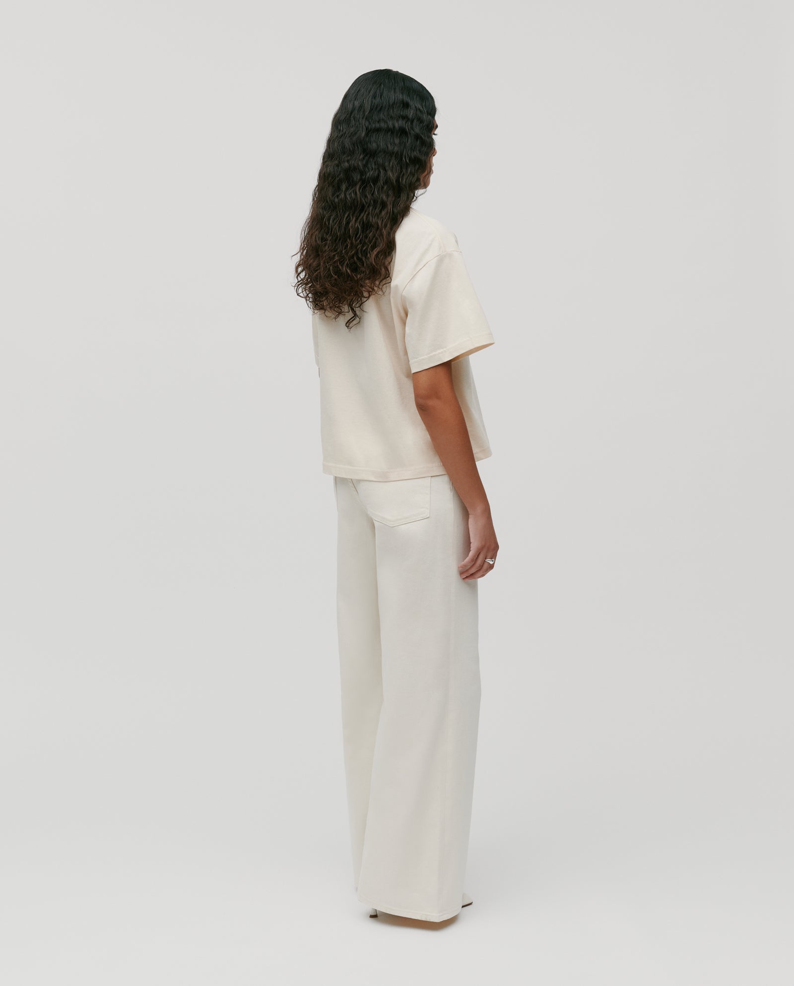 Person with long curly hair wears the IVY OAK KYLA Top—a must-have loose cream boxy T-shirt—paired with wide-leg pants, standing with their back to the camera.