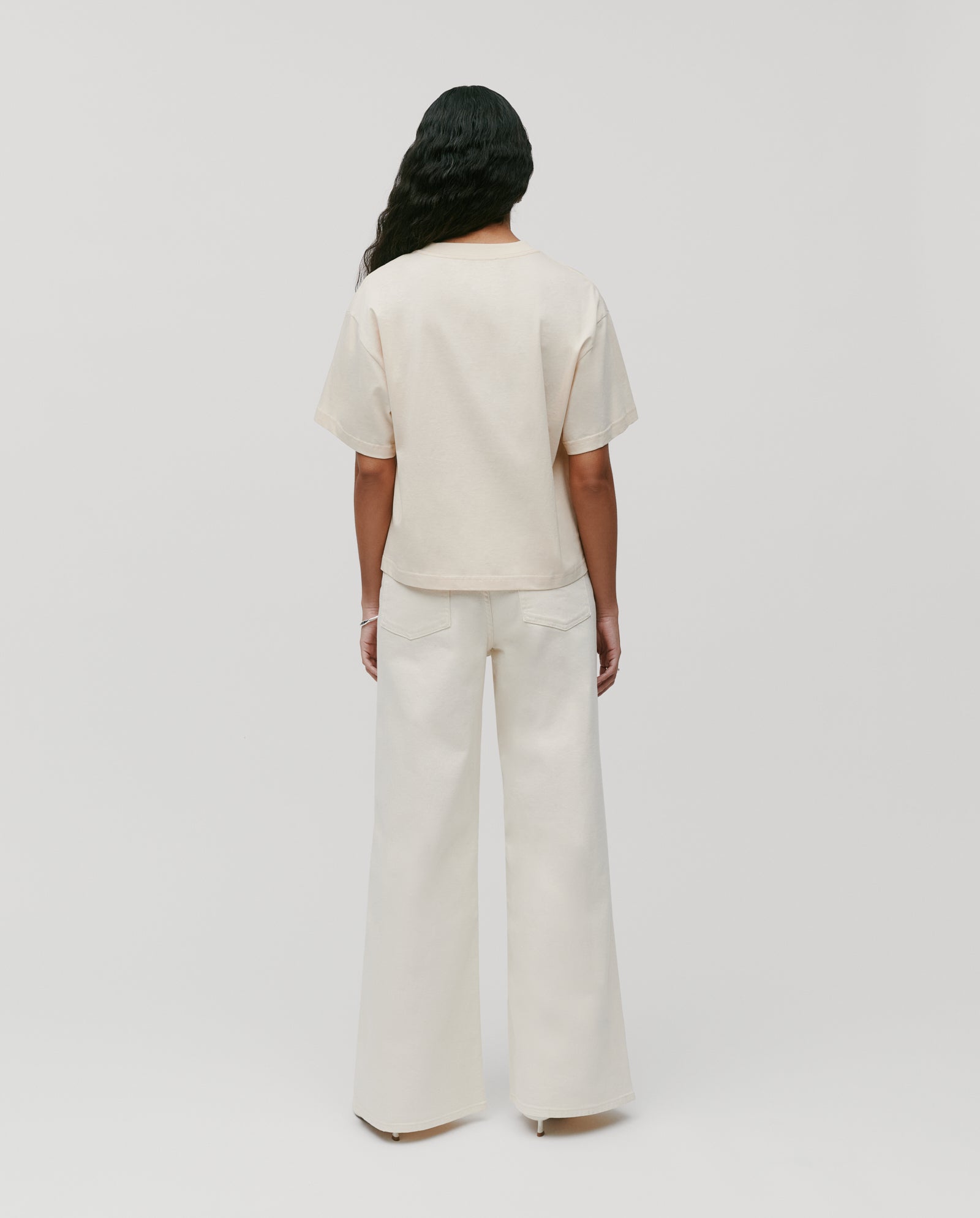 A person stands with their back to the camera, wearing the IVY OAK KYLA Top—a must-have boxy T-shirt in off-white, crafted from soft organic cotton.