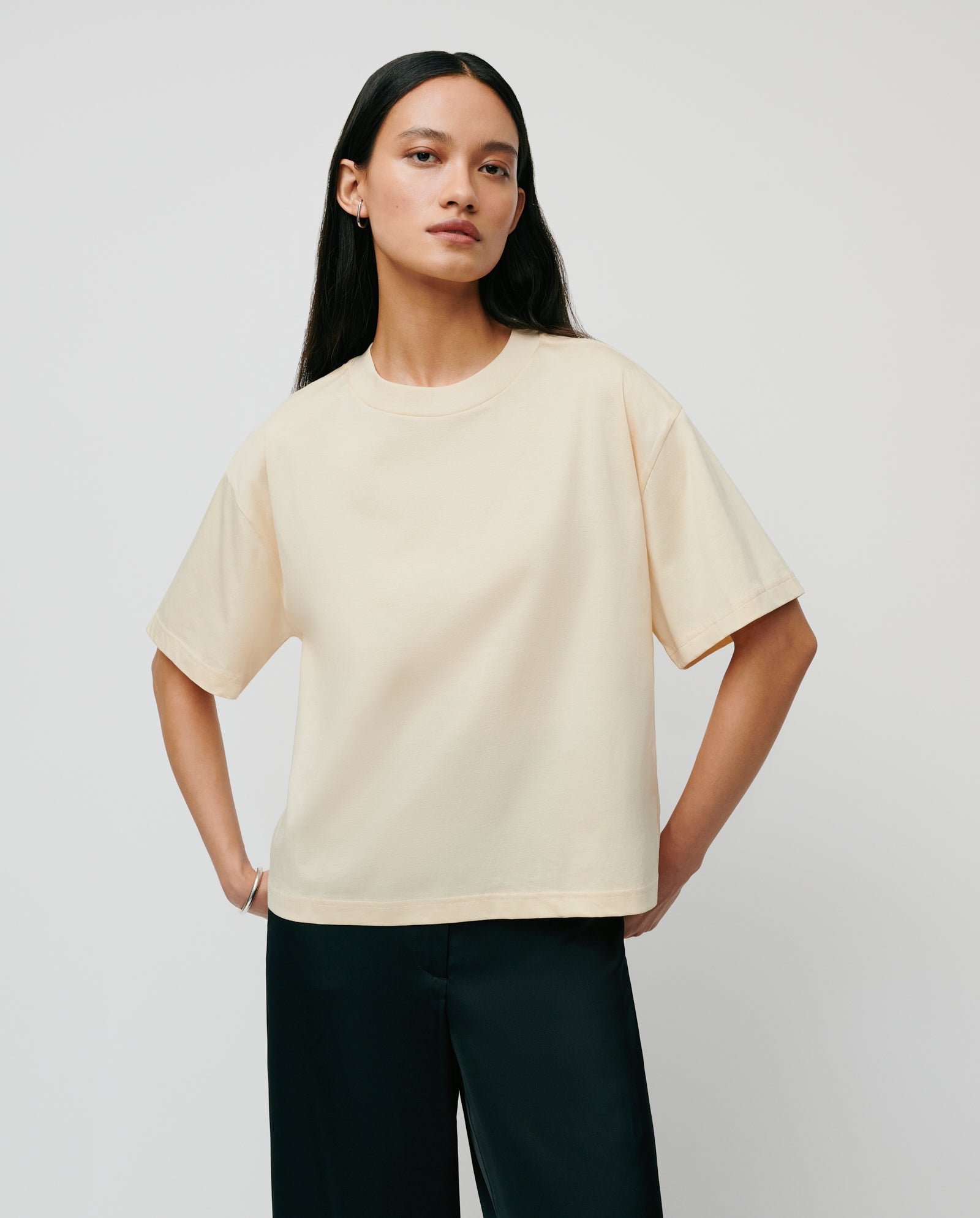 A woman stands with hands on hips against a plain background, wearing the IVY OAK KYLA Top in cream organic cotton and black pants—a basic must-have for any wardrobe.