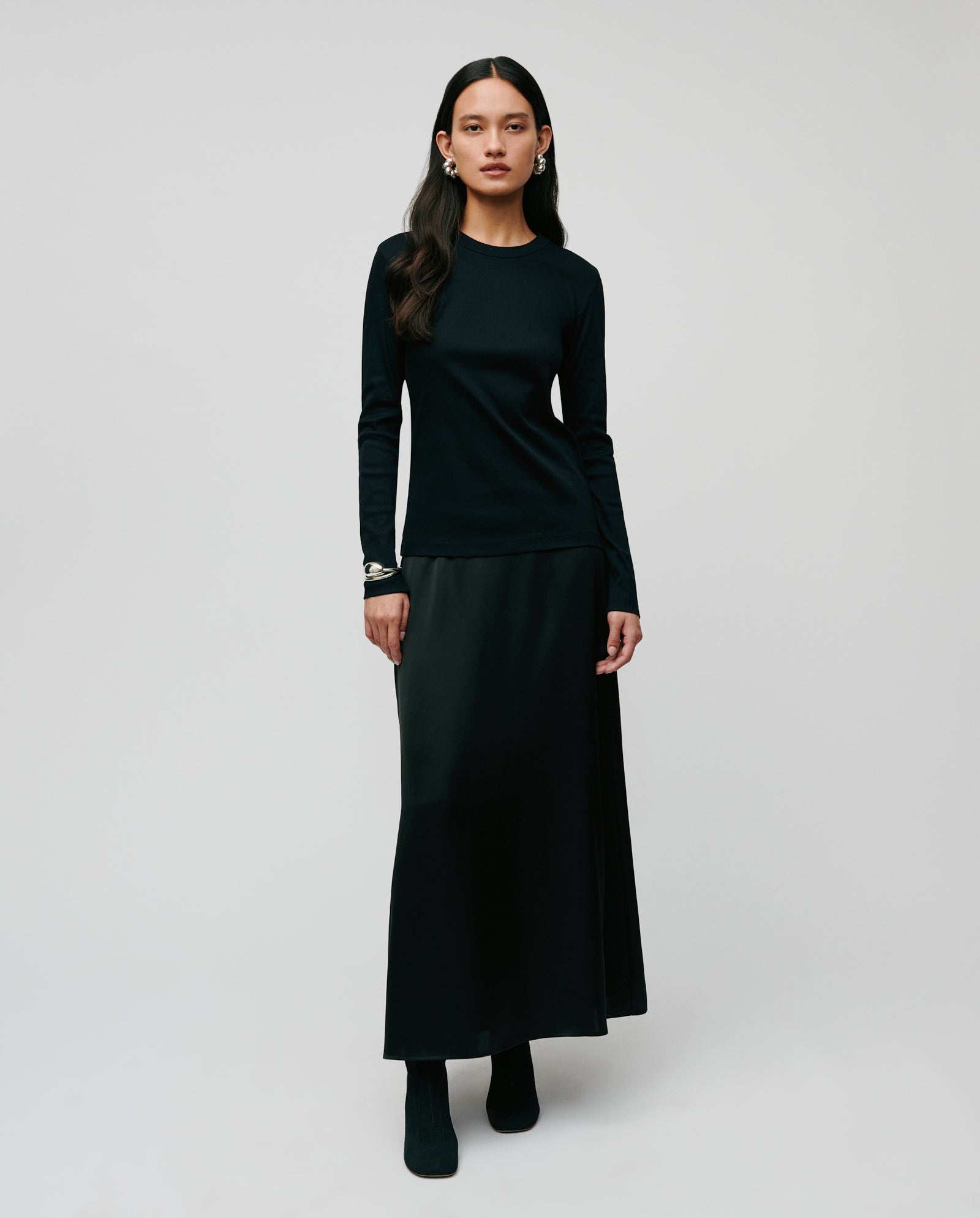 A woman in a long black skirt, comfortable IVY OAK KOLLINA Top made of Organic Cotton, and boots stands against a plain, light background.
