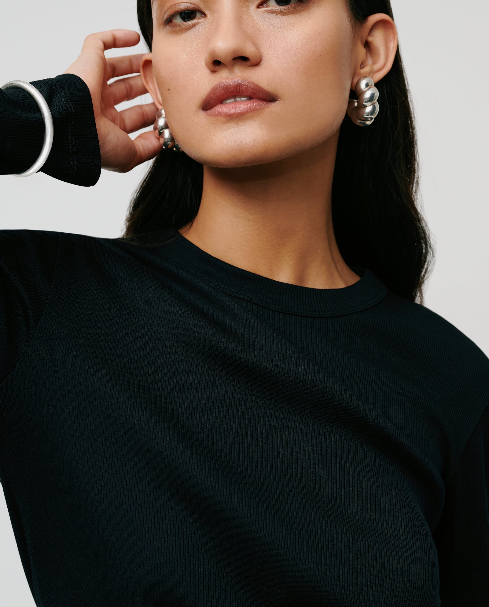 Woman in a comfortable black IVY OAK KOLLINA Top made from organic cotton, paired with silver hoop earrings and a silver bracelet, touching her ear.
