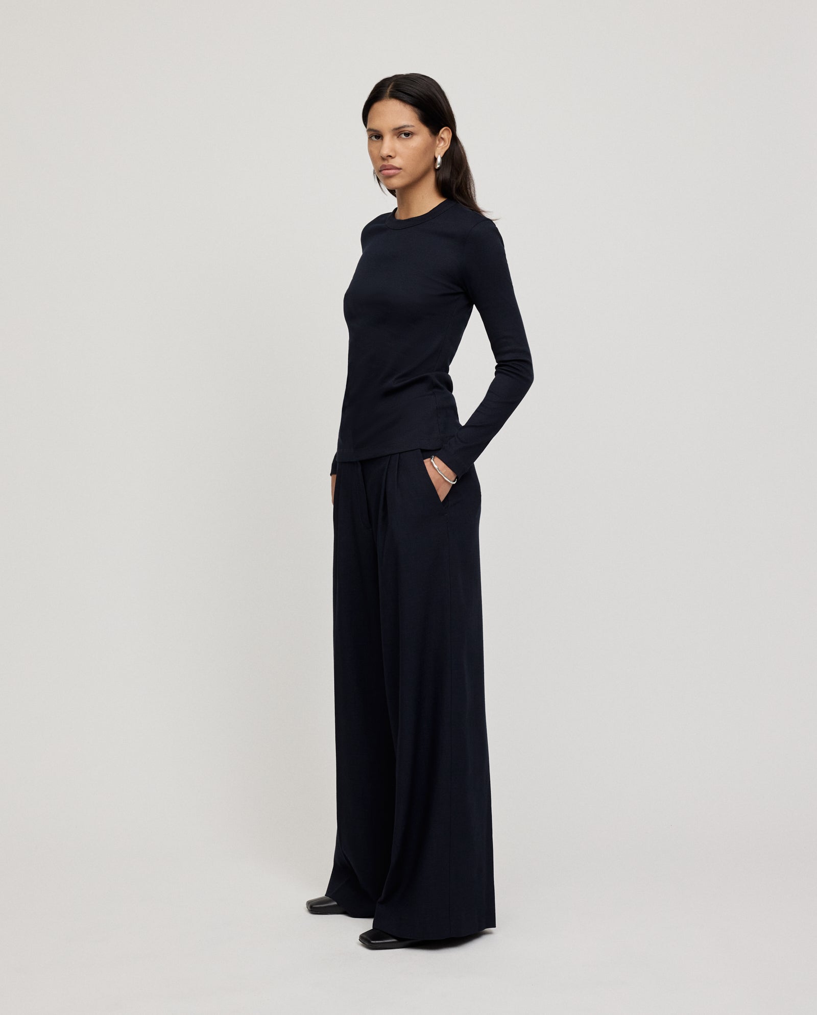 A woman wears the IVY OAK KOLLINA Top, a comfortable black long-sleeve round neck, paired with wide-leg pants as she stands against a plain light background with her hands in her pockets.