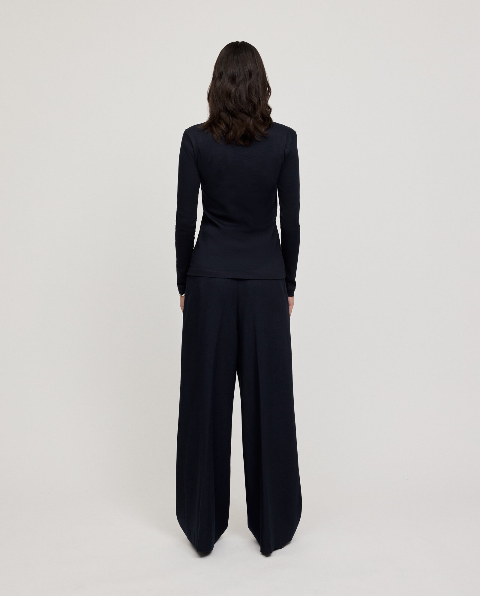 A person with wavy brown hair, standing facing away, wears the IVY OAK KOLLINA Top—a black, round neck piece made from organic cotton—paired with wide-leg pants.