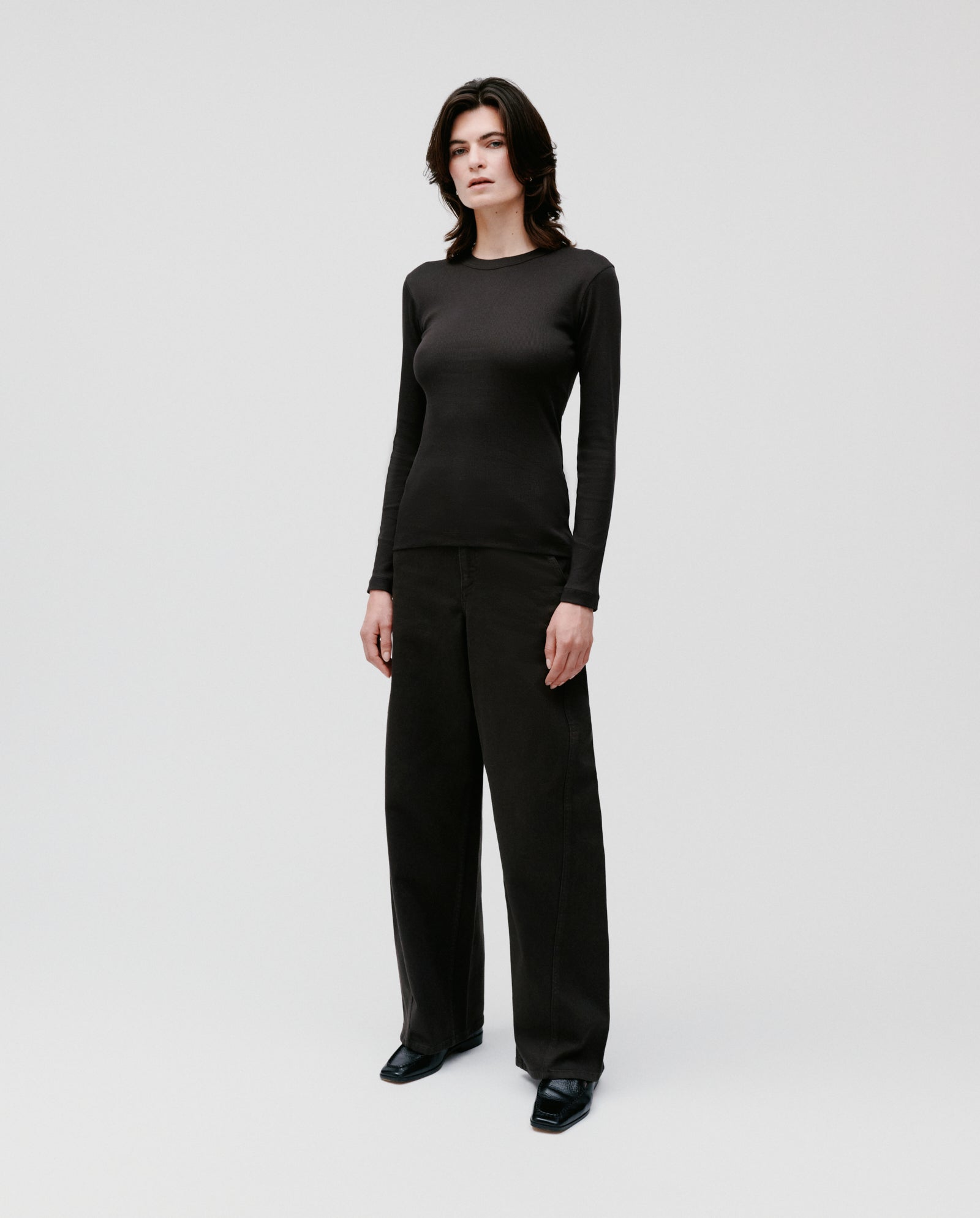 A person wears the IVY OAK KOLLINA Top— a comfortable, black, long-sleeve round neck made from organic cotton—paired with wide-leg black pants, standing against a plain white background.