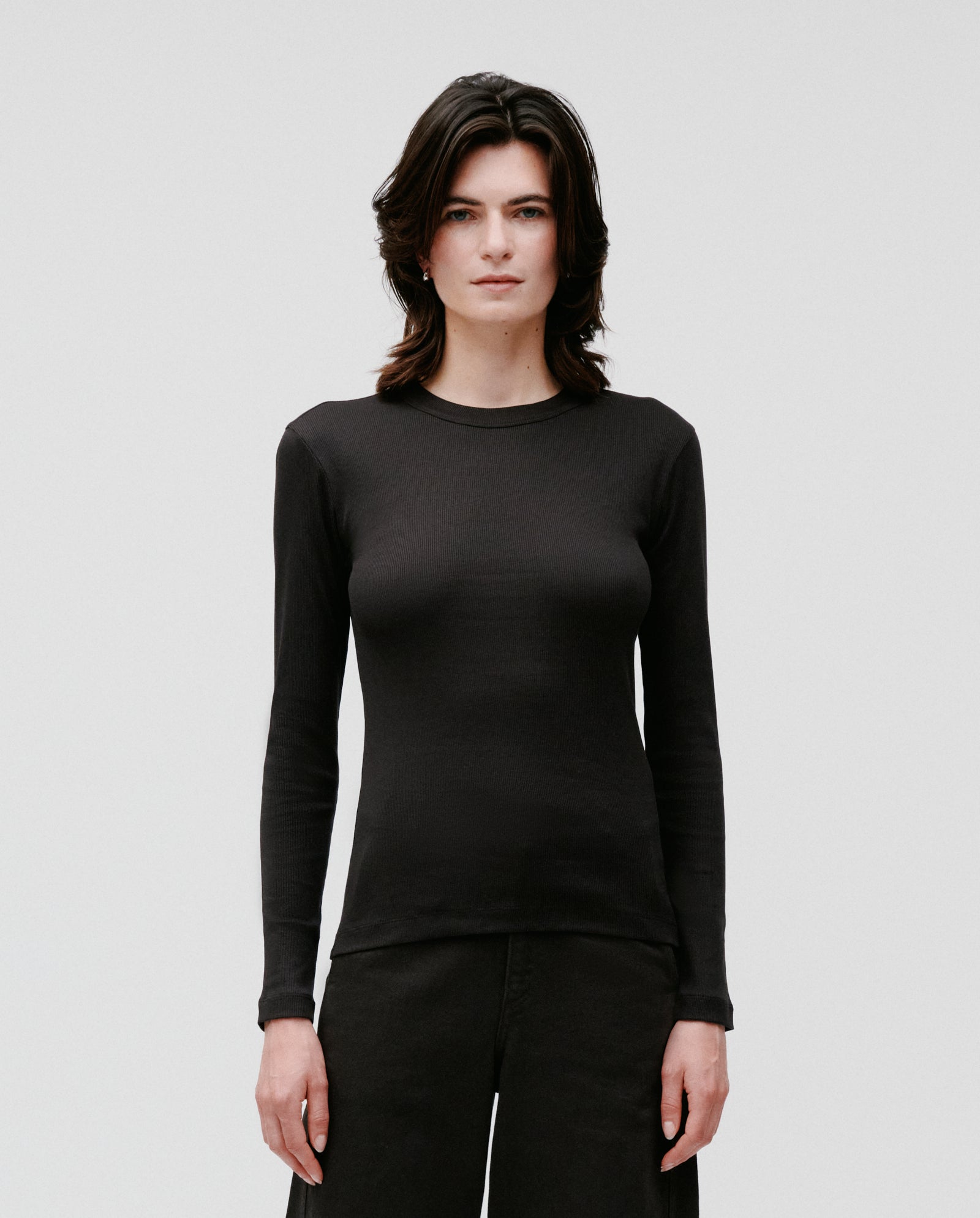 A person wears the IVY OAK KOLLINA Top—a fitted, long-sleeve black organic cotton shirt with a round neck—paired with black pants, standing against a plain white background.