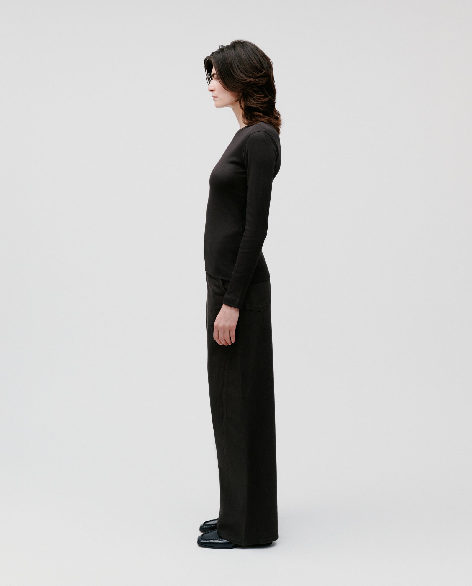 A woman stands in profile against a plain light background wearing the IVY OAK KOLLINA Top, a comfortable black long-sleeve with a round neck, paired with wide-leg pants.