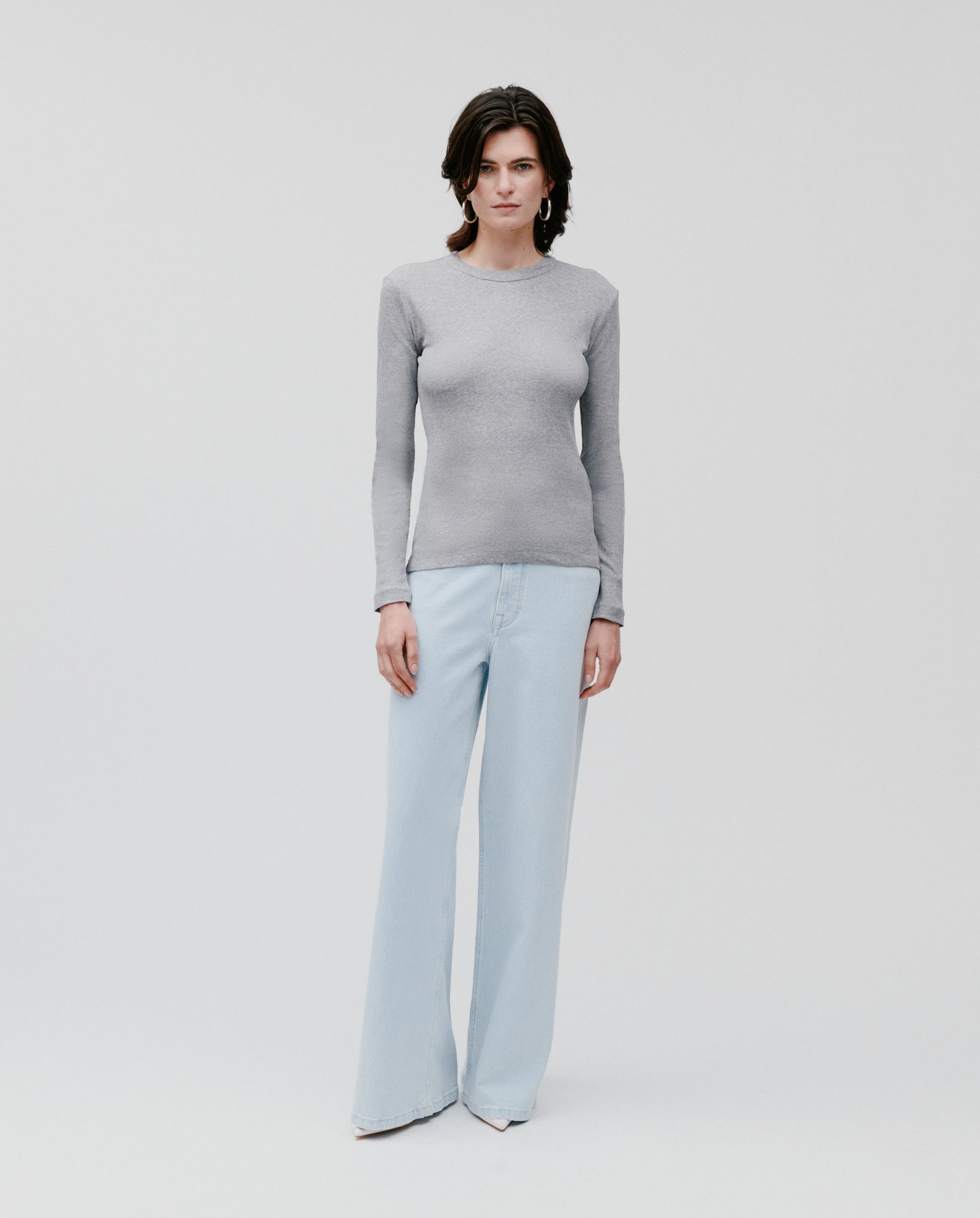 A woman models the IVY OAK KOLLINA Top, a gray, long-sleeve, round-neck shirt made from soft organic cotton, paired with light blue wide-leg jeans against a plain white background.