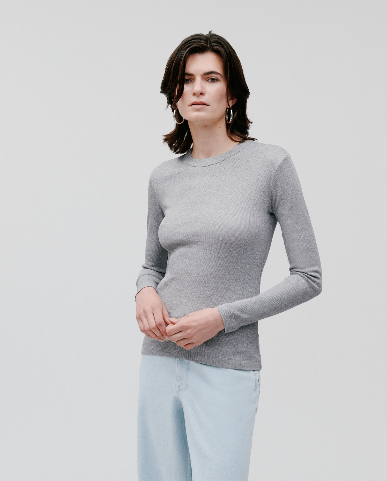 A person with medium-length brown hair wears the IVY OAK KOLLINA Top, a comfortable round neck gray long-sleeve, paired with light blue jeans, standing against a plain background.