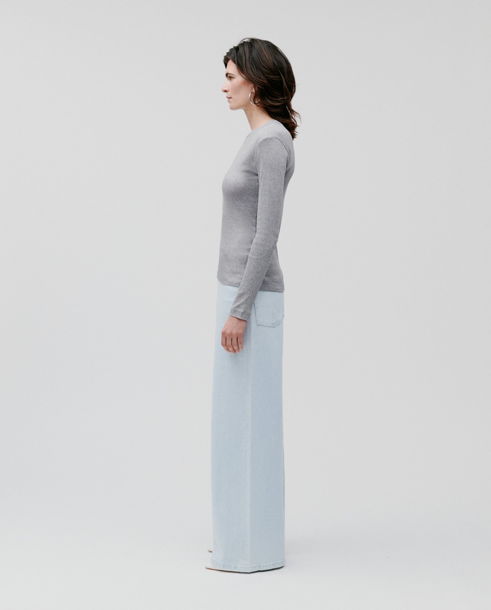 A woman stands in profile wearing the IVY OAK KOLLINA Top, a comfortable organic cotton grey round-neck long-sleeve, paired with light blue wide-leg pants against a plain backdrop.