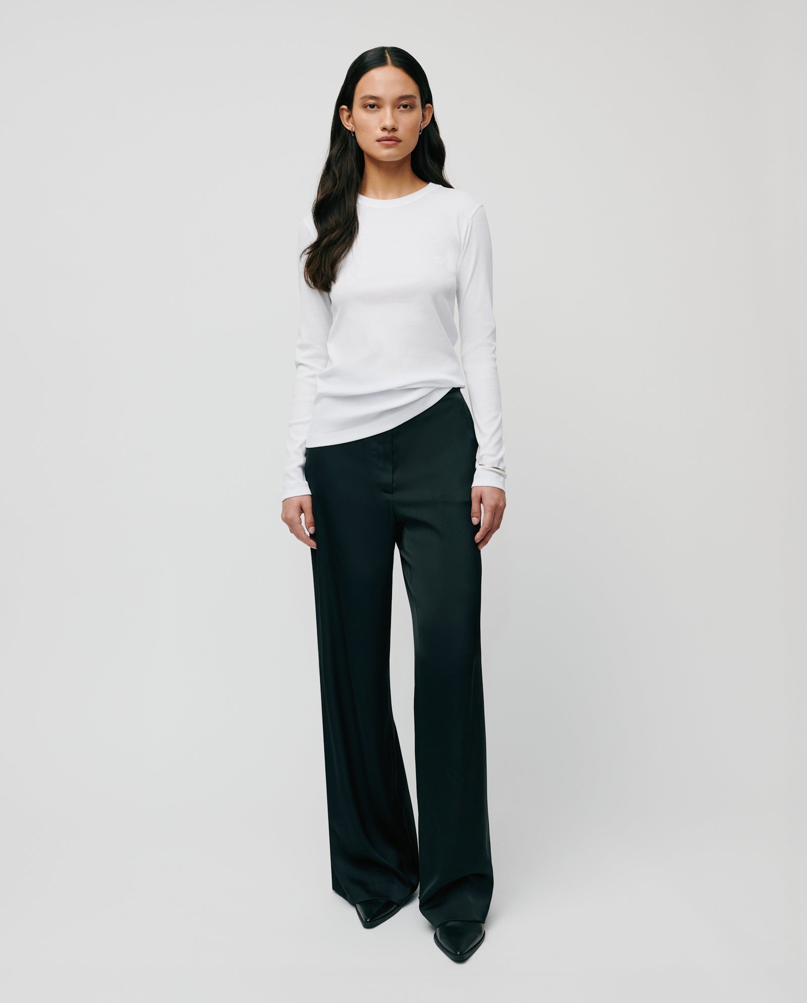A woman stands against a plain light background, wearing the IVY OAK KOLLINA Top in white organic cotton and black wide-leg trousers.