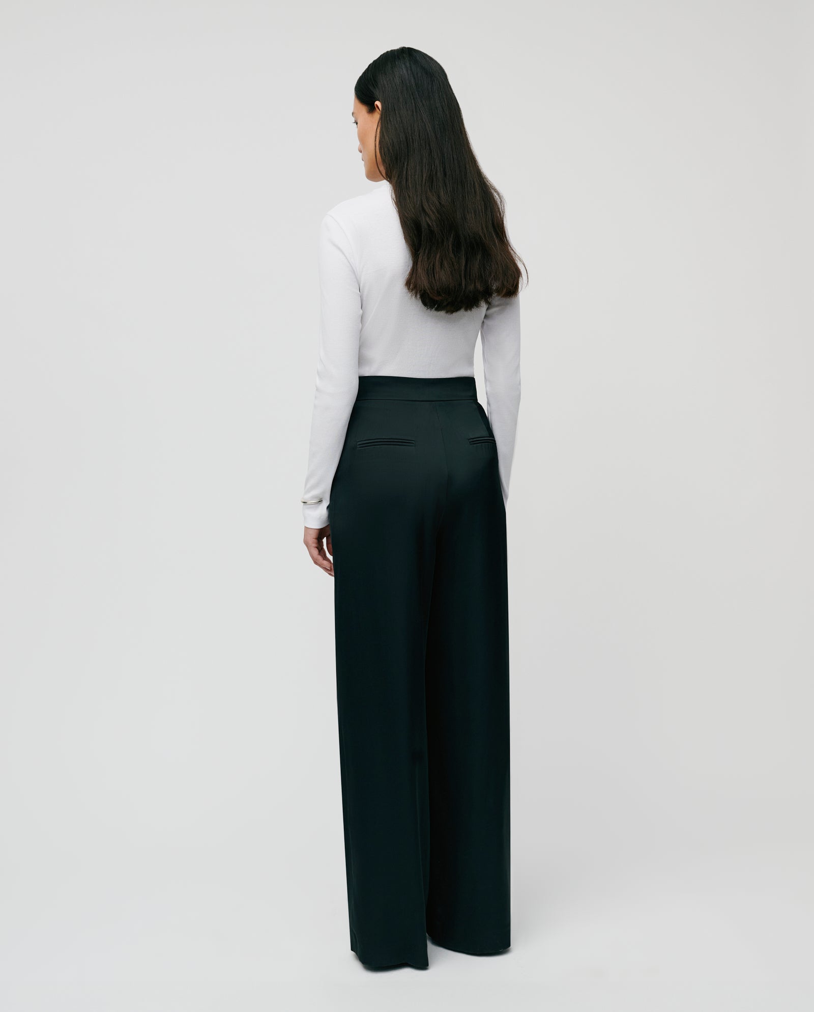 A woman stands with her back to the camera, wearing the IVY OAK KOLLINA Top in organic cotton and black wide-leg pants.