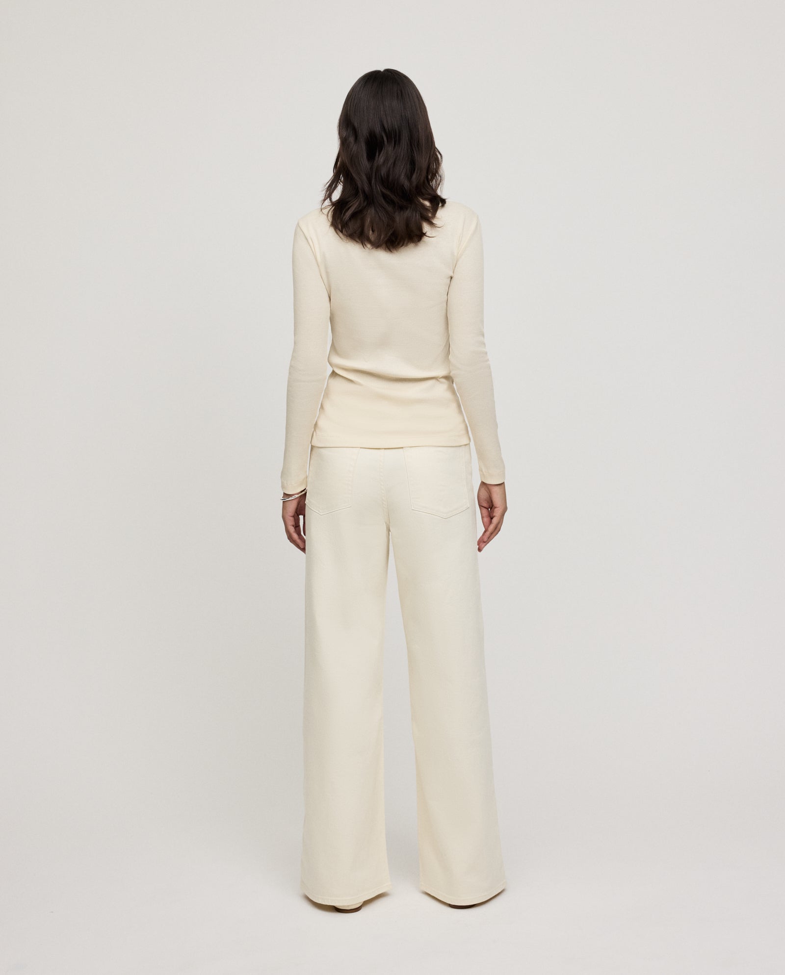 A woman with wavy dark hair, wearing the IVY OAK KOLLINA Top in organic cotton and wide-leg pants, stands facing away.
