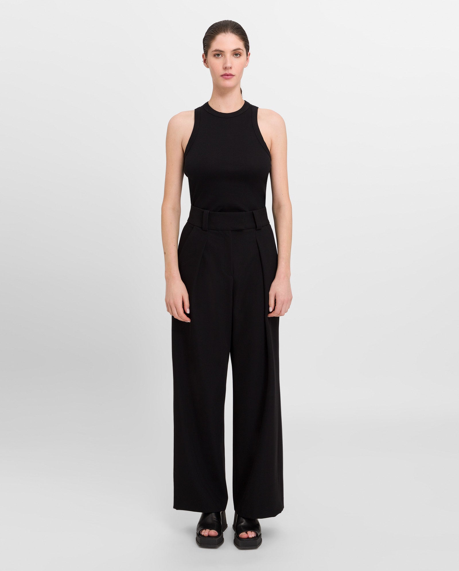 A woman stands against a plain background wearing the KLARA ANN Top by IVY OAK, paired with wide-leg black pants and black platform sandals.