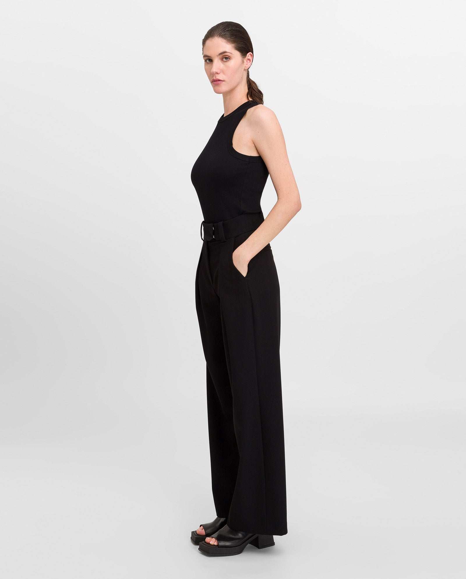 A woman in profile wears the IVY OAK KLARA ANN Top in black organic cotton, styled with wide-leg pants and heeled sandals against a plain background.