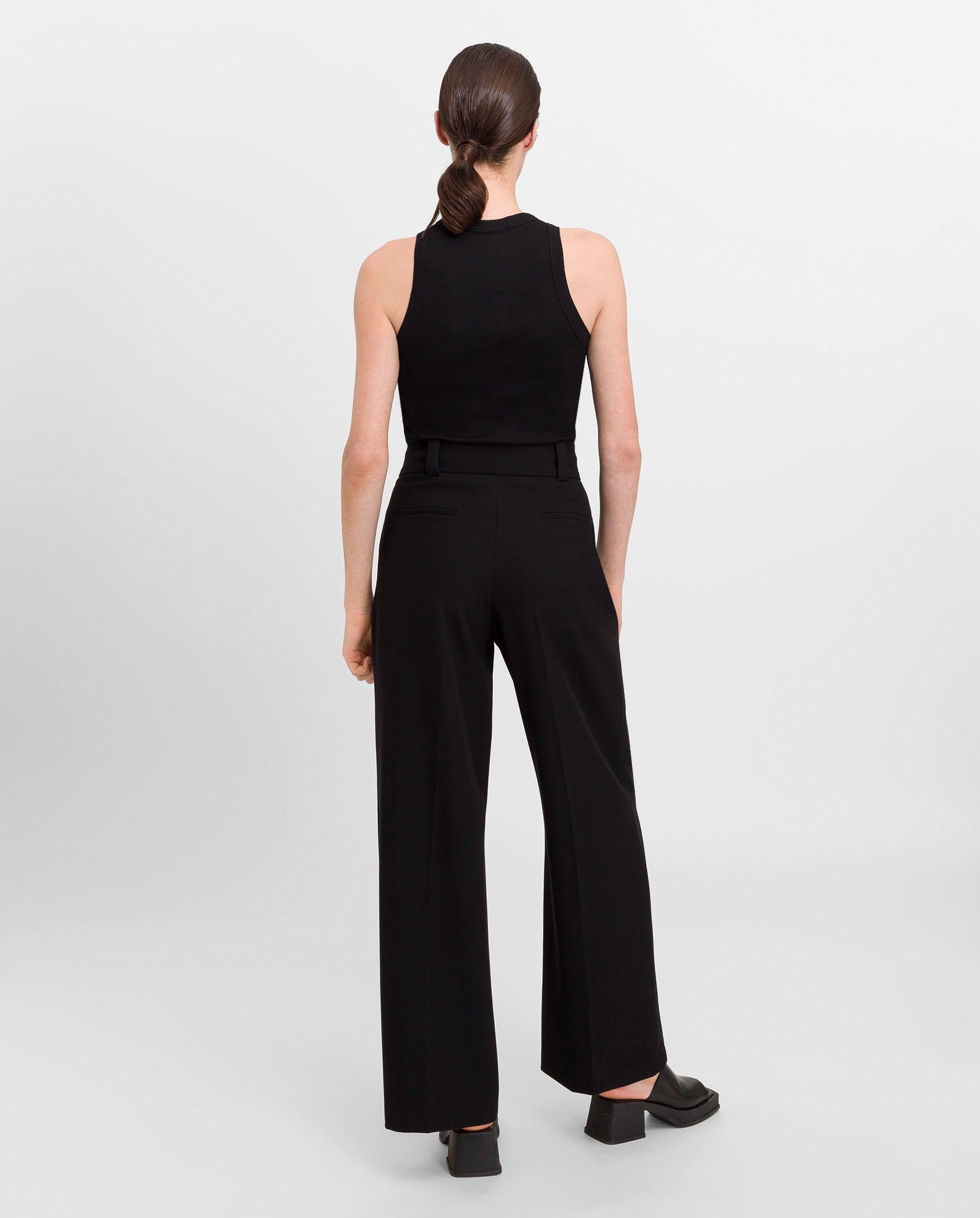 Woman with brown hair in a low ponytail, wearing the IVY OAK KLARA ANN Top and wide-leg black pants, facing away.