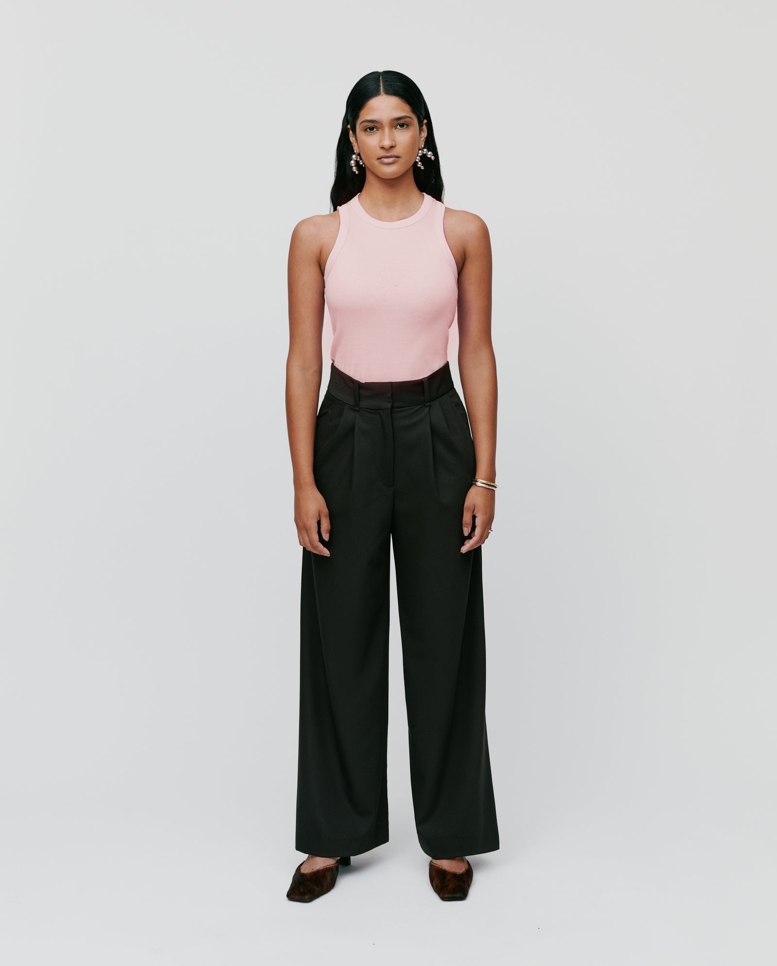 A woman stands facing forward against a plain background, wearing the IVY OAK KLARA ANN Top in pink paired with black wide-leg trousers for a comfortable look.