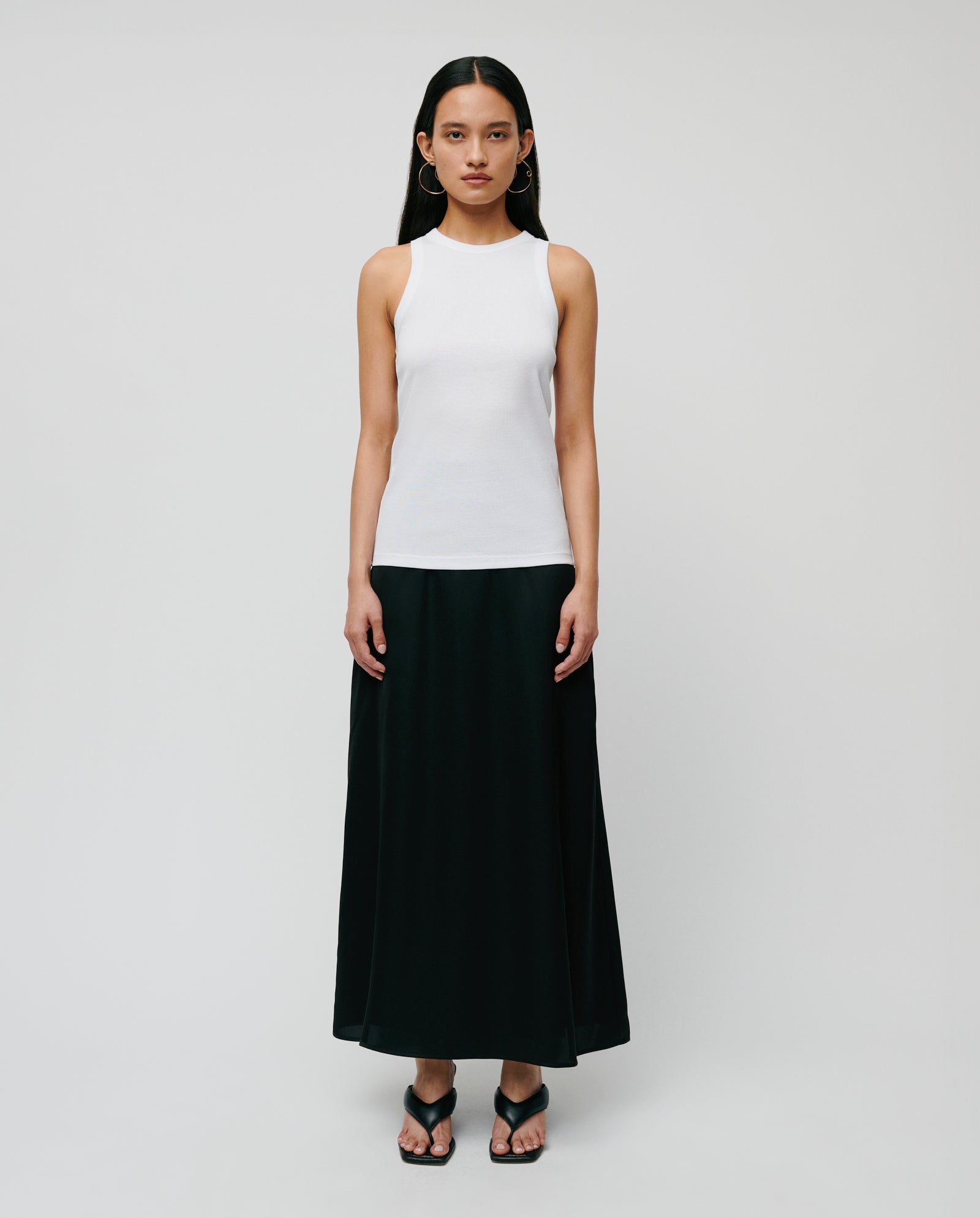 A woman wears the IVY OAK KLARA ANN Top, an organic cotton tank, paired with a long black skirt and black sandals against a plain background.