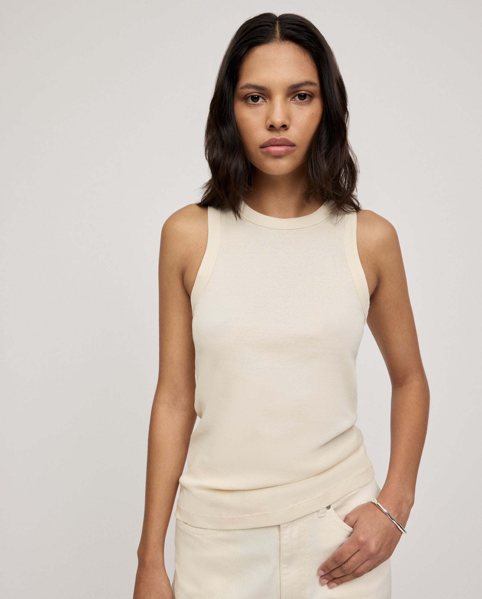 A woman with shoulder-length dark hair wears the KLARA ANN Top by IVY OAK—a comfortable beige tank made from Organic Cotton—paired with light pants, standing against a plain background.