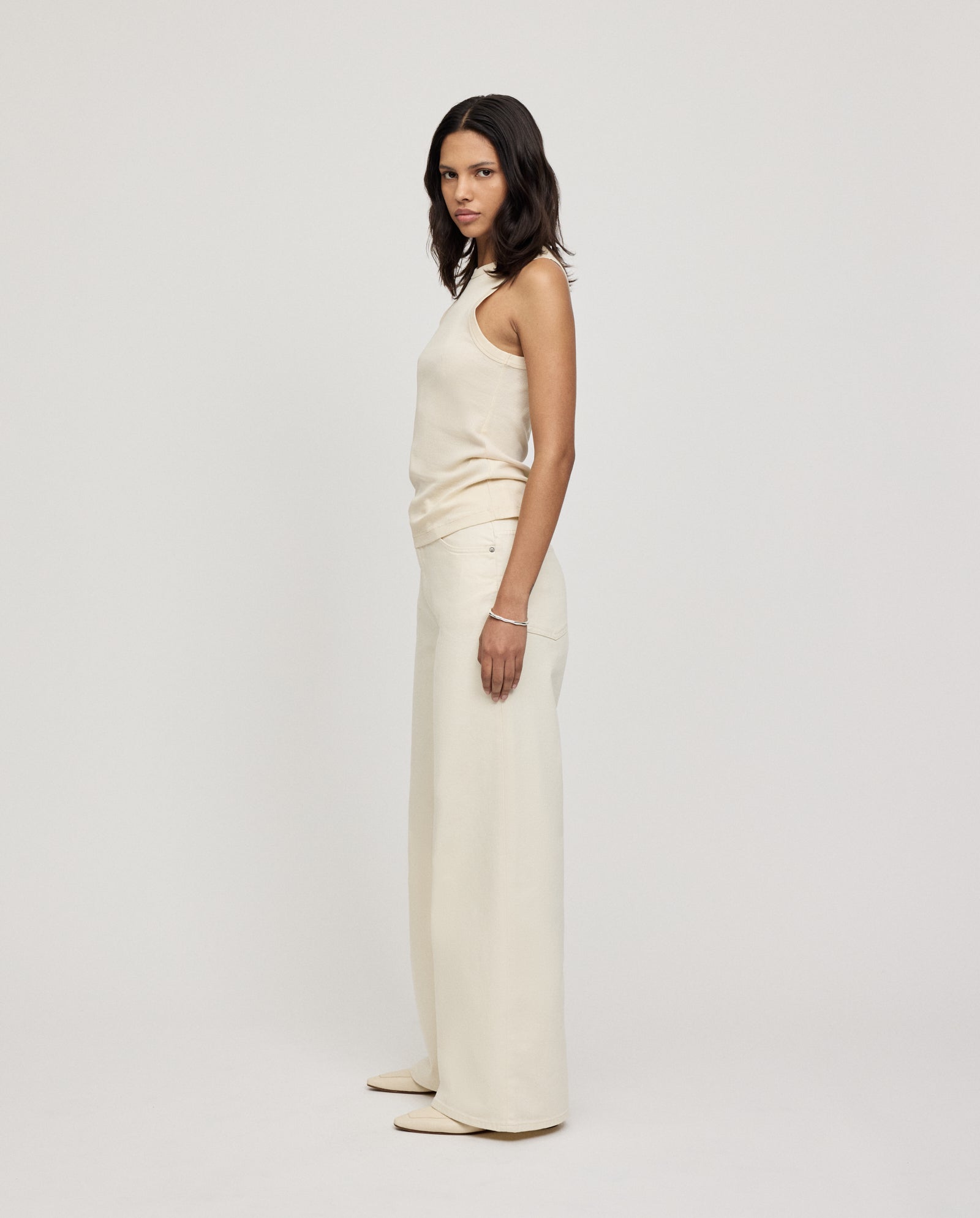 A woman wearing the IVY OAK KLARA ANN Top, a comfortable cream tank, pairs it with wide-leg pants as she stands sideways against a plain light background.