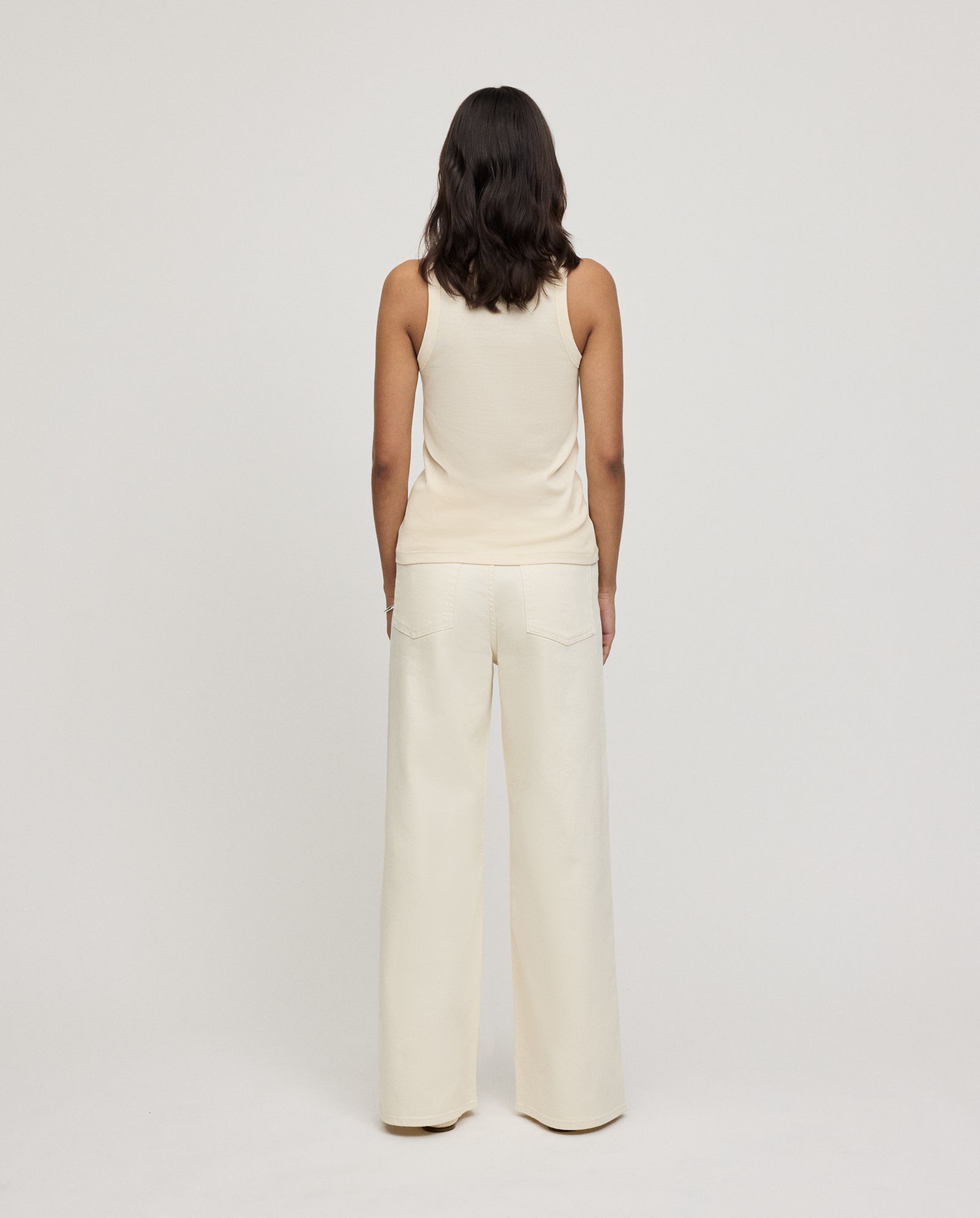 Woman with shoulder-length dark hair wears the IVY OAK KLARA ANN Top—an off-white, organic cotton tank—paired with wide-leg pants, both crafted from organic cotton, facing away.