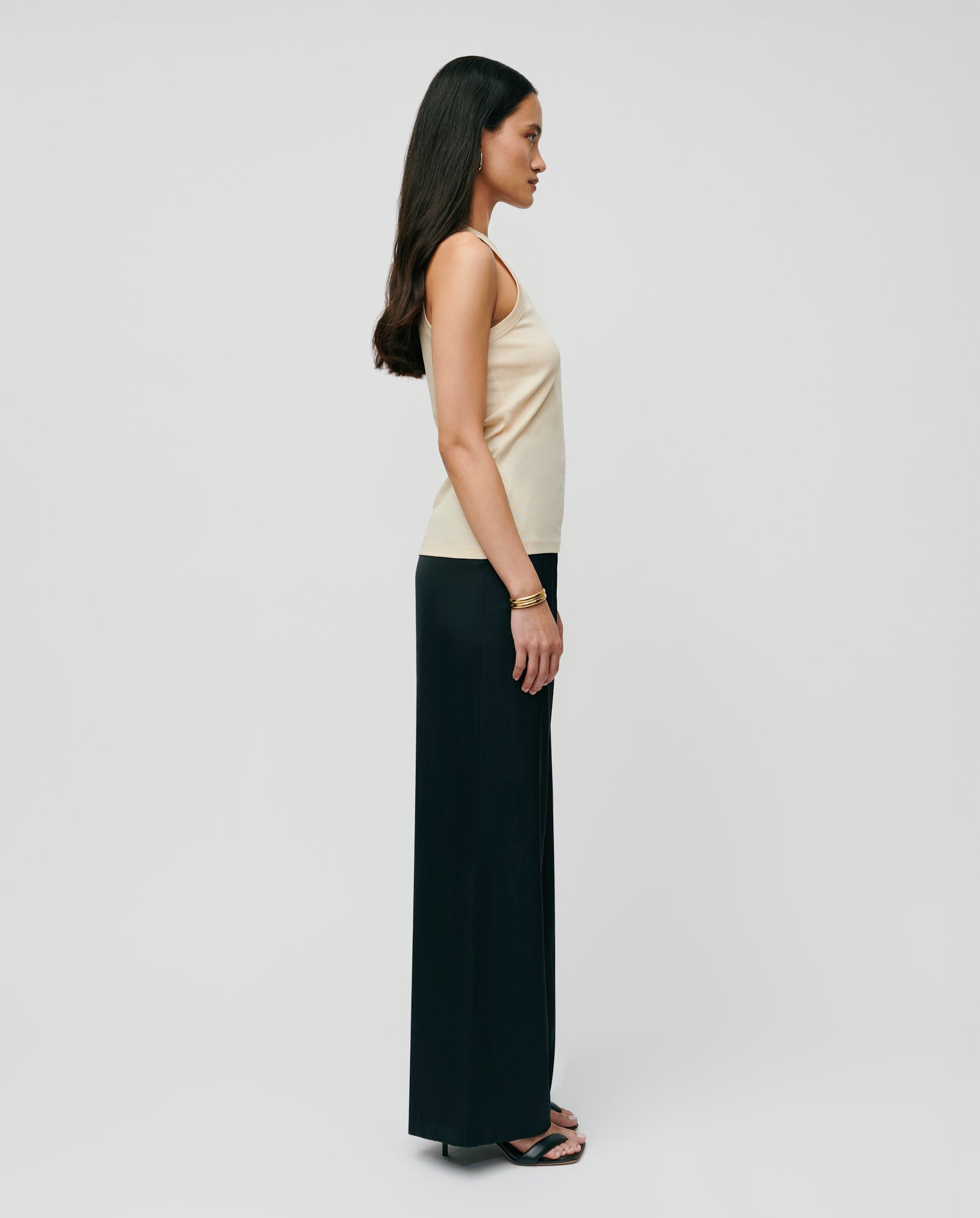 A woman stands in profile against a plain white background, wearing the KLARA ANN Top by IVY OAK—an organic cotton beige tank—and black wide-leg pants.