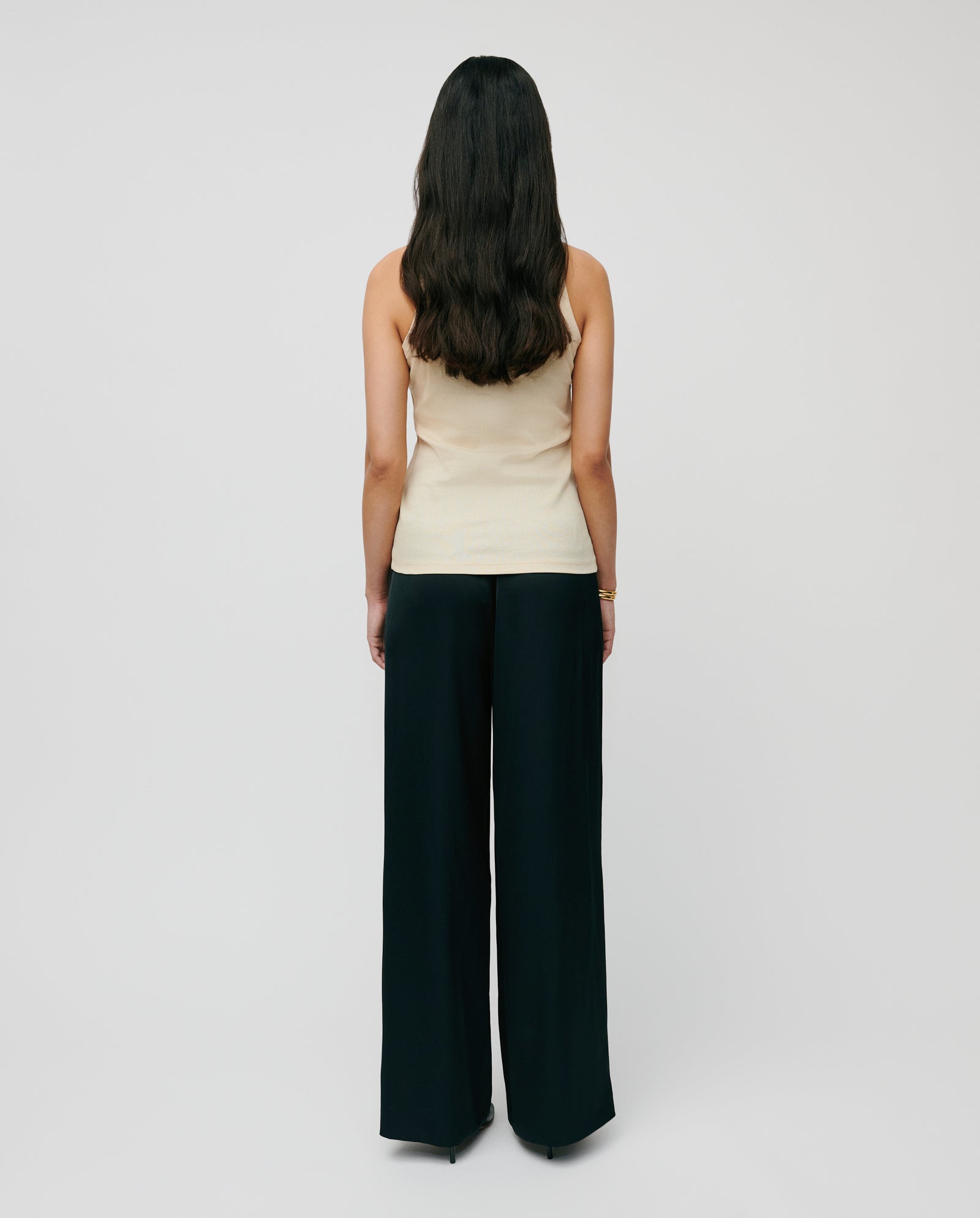 A woman with long dark hair faces away, wearing the IVY OAK KLARA ANN beige organic cotton tank top and black wide-leg pants.